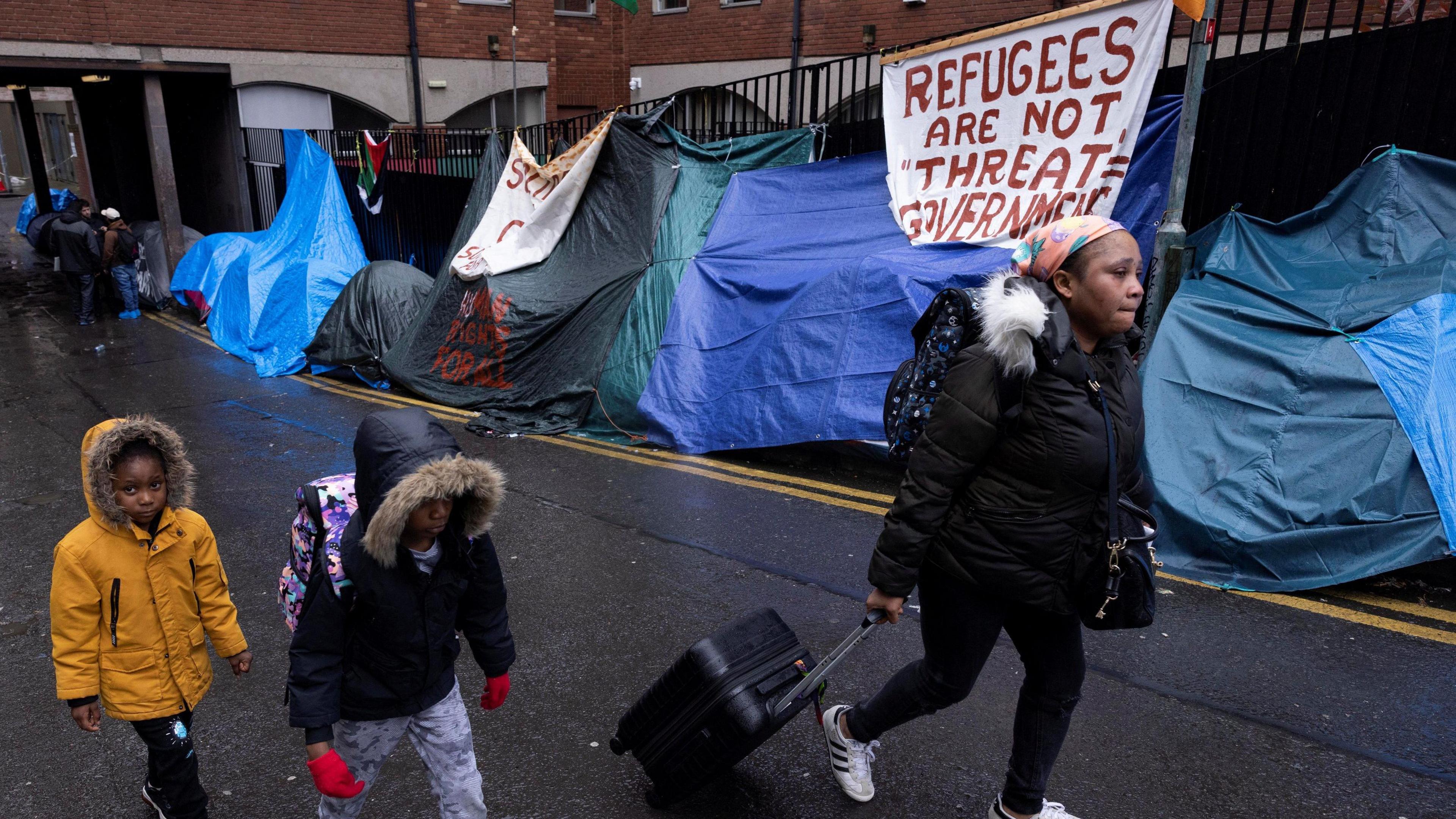 Three asylum seekers (two small children and a young girl) walk past tents outside the International Protection Office (IPO). A banner saying "Refugees are not 'threat' government"