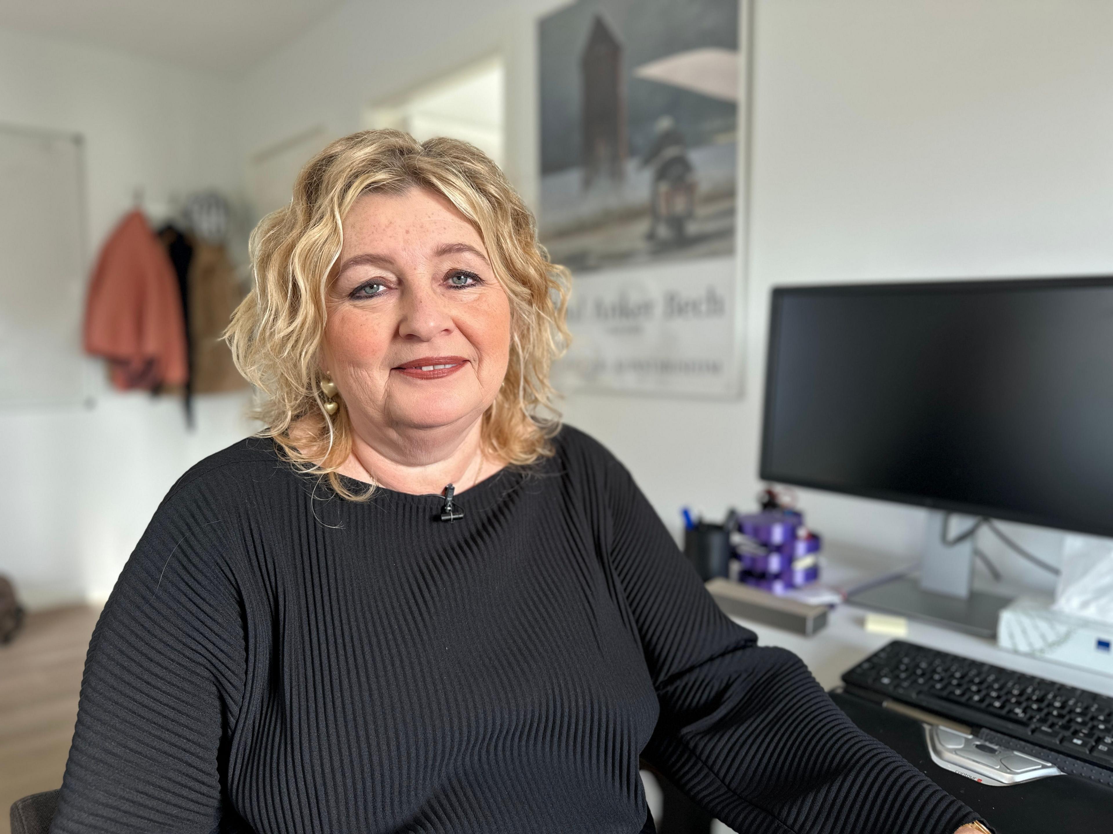 A woman with shoulder-length wavy blonde hair, Tordis Jacobsen, looks at the camera. She is wearing a black jumper and is sitting next to a desk with a computer in an office.