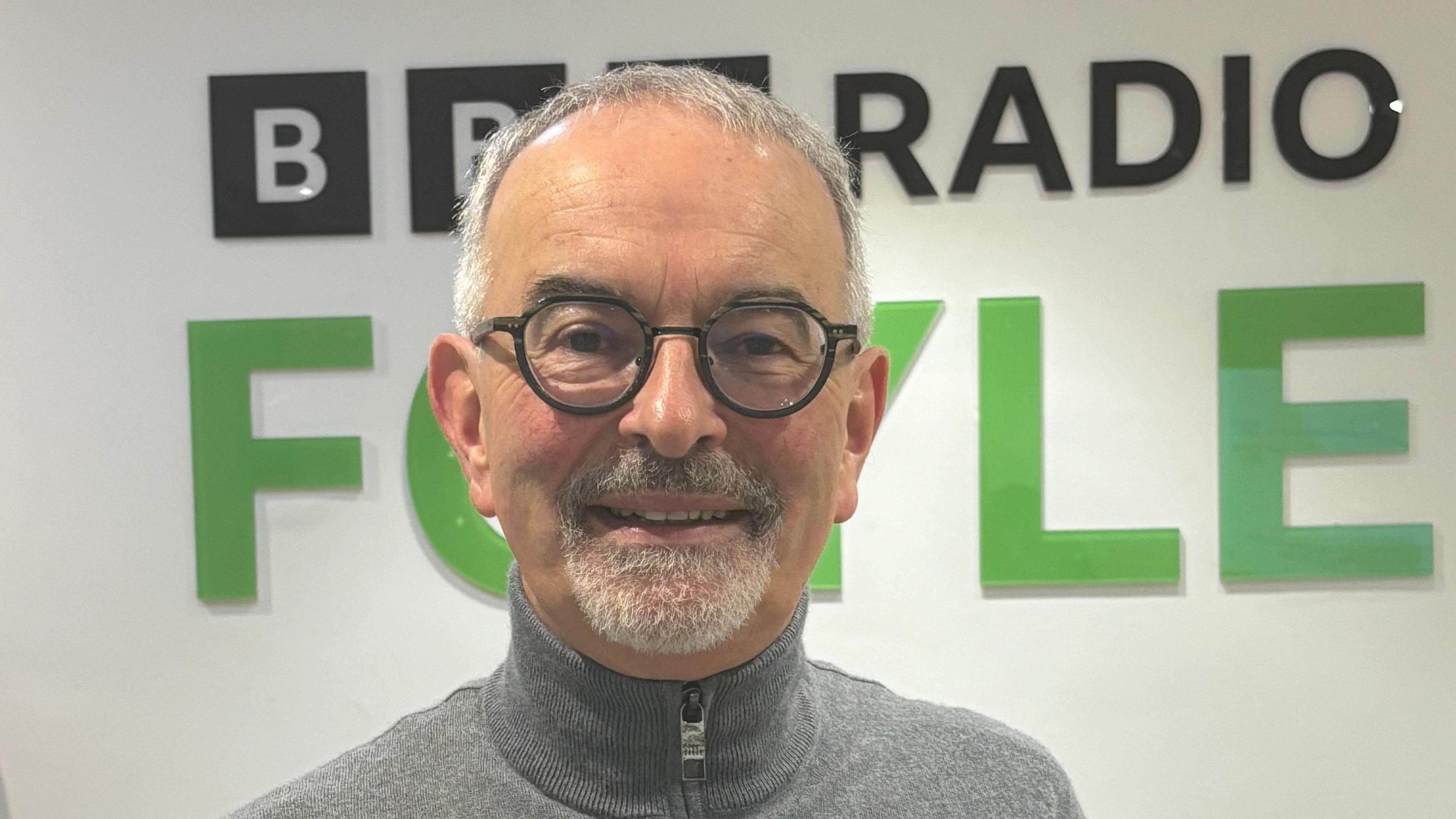 Dónal Doherty pictured wearing dark-rimmed glasses and in a grey zip-up sweater. He is standing in front of BBC Radio Foyle sign.