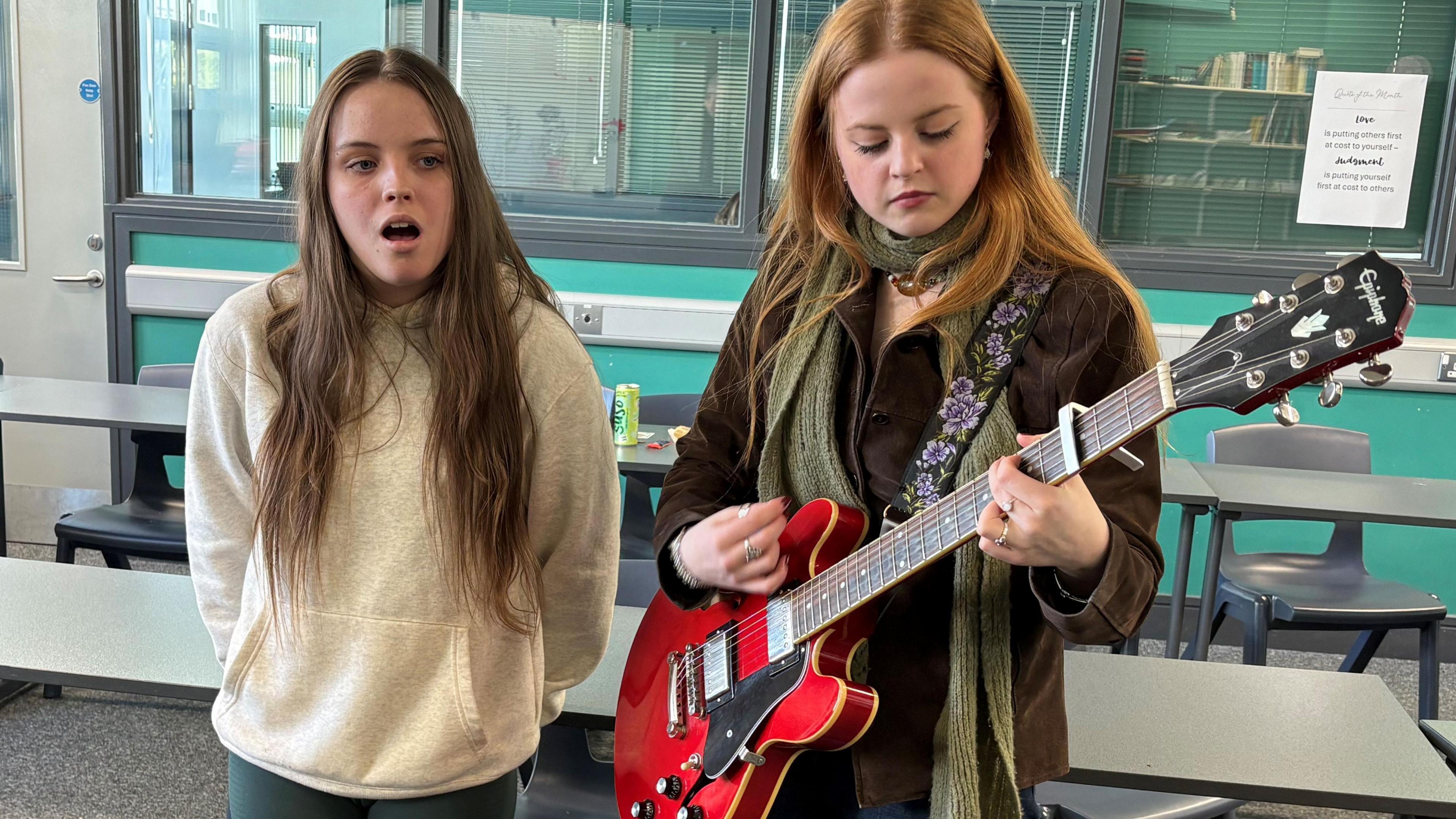 Ebony (left) is singing and Olivia (right) is playing a red guitar. Ebony is wearing a white hoody and green leggings and Olivia is wearing a brown jacket and green scarf. There are tables and chairs in the classroom behind them. 