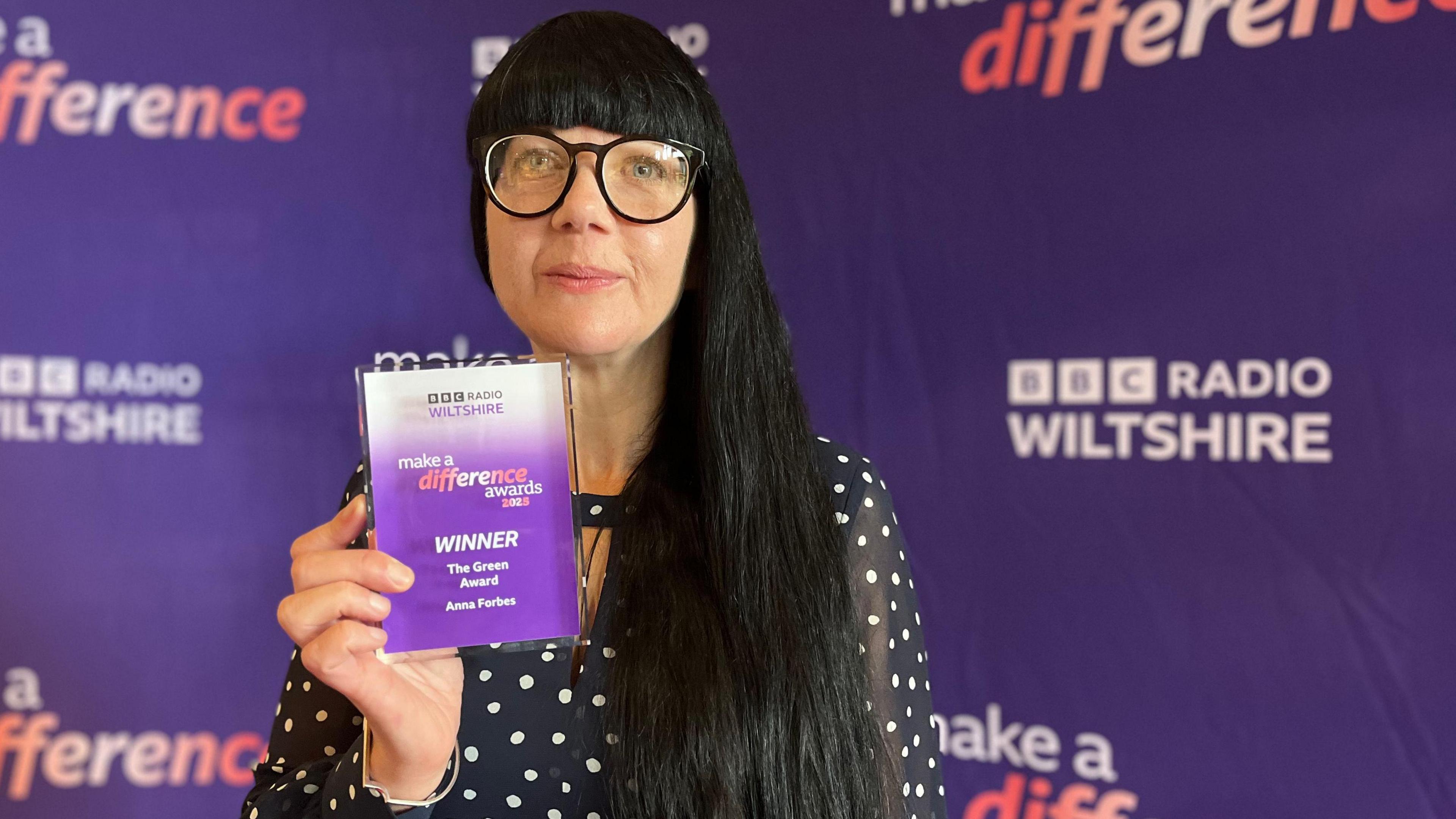 Anna Forbes looks at the camera holding her award. She wears glasses and polka dot dress.
Behind her is a purple banner saying BBC Radio Wiltshire and Make a Difference.