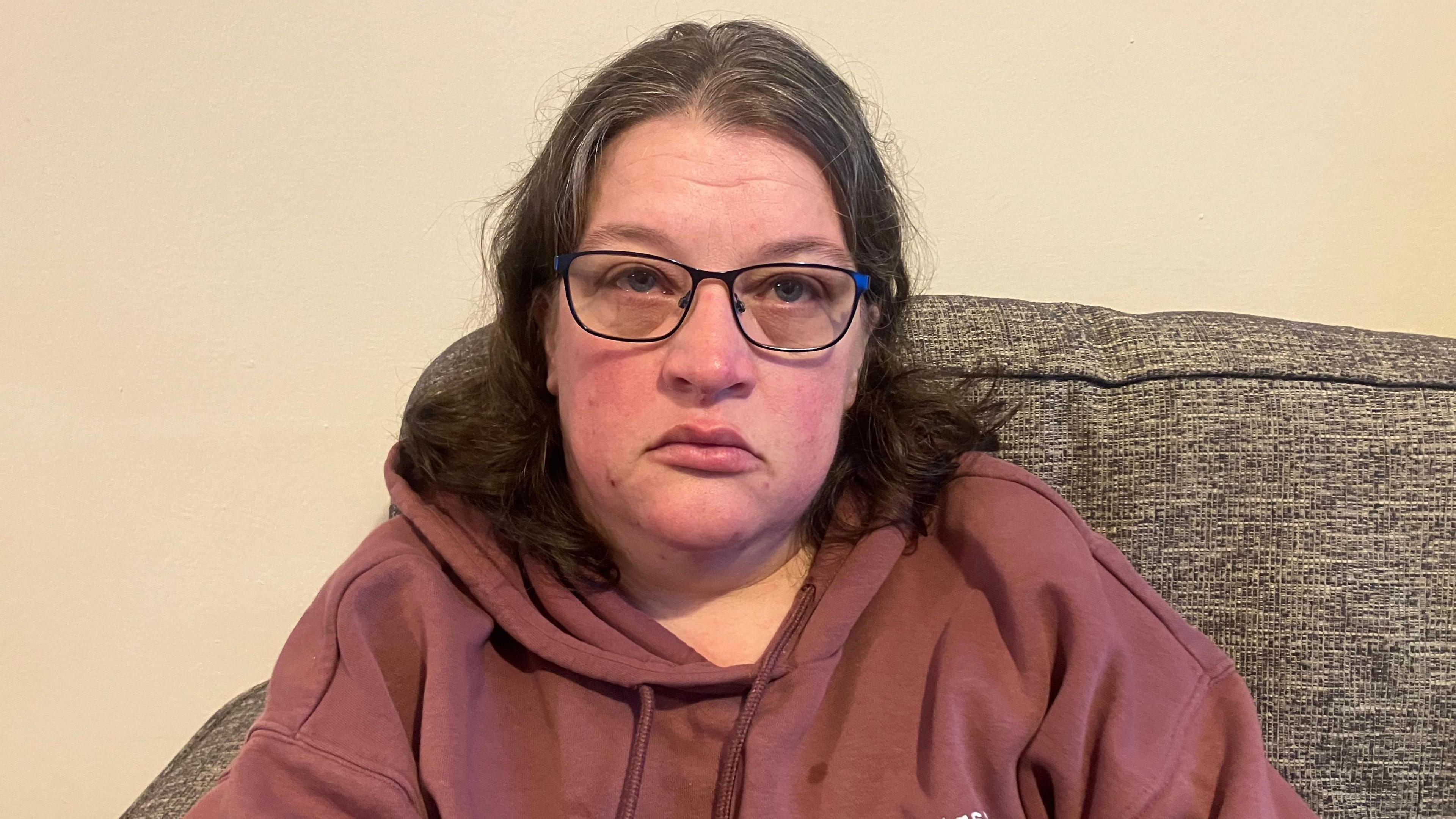Abbie sitting on the sofa. She is wearing a brown hoodie and dark square glasses, with her dark hair sitting at her shoulders. She is straight faced and looking at the camera. 