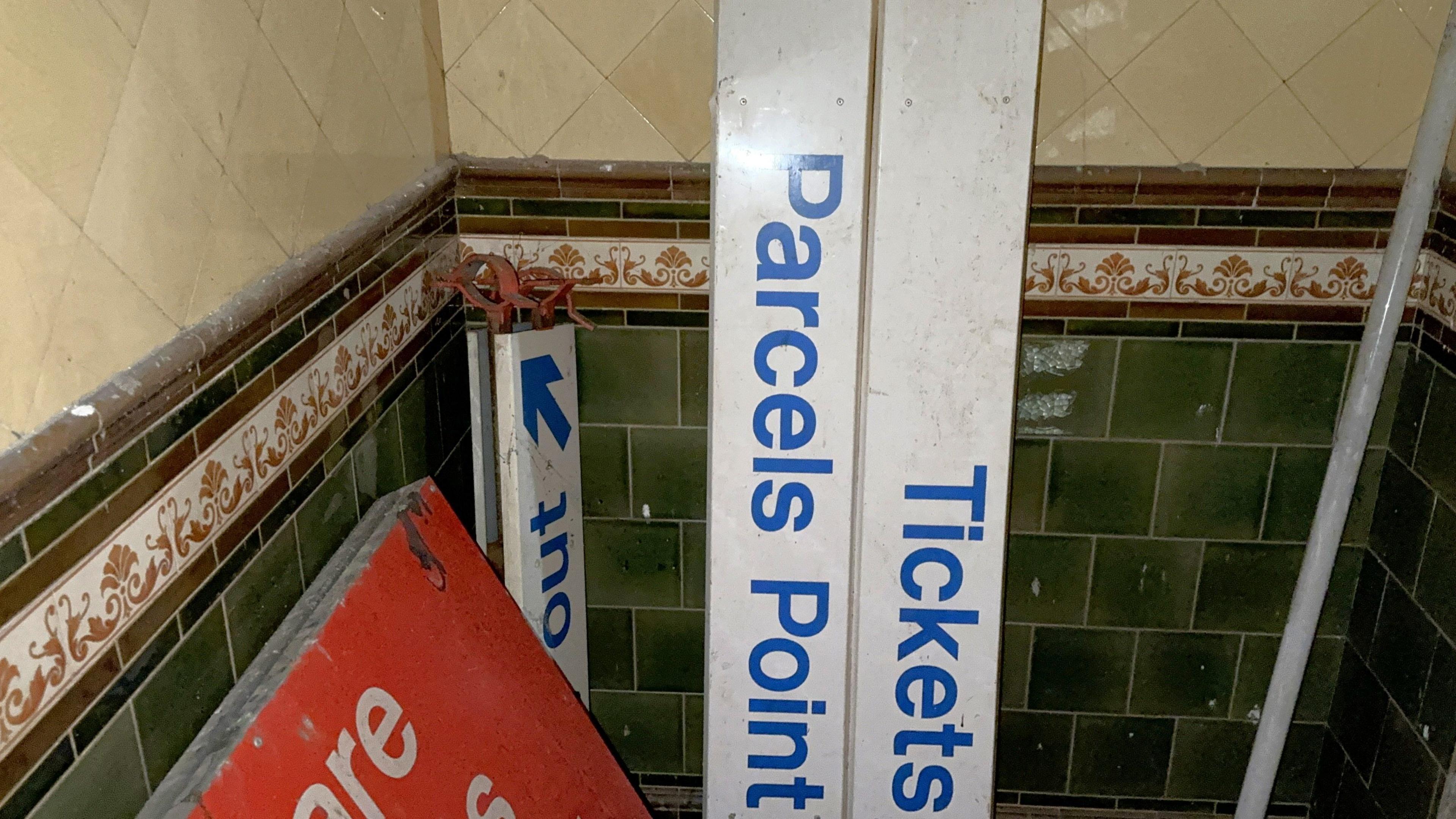 Old signs propped up in the corner of a dark underground room. The walls are covered in yellowing square tiles on the top and dark green tiles on the bottom, separated by a brown patterned border. 