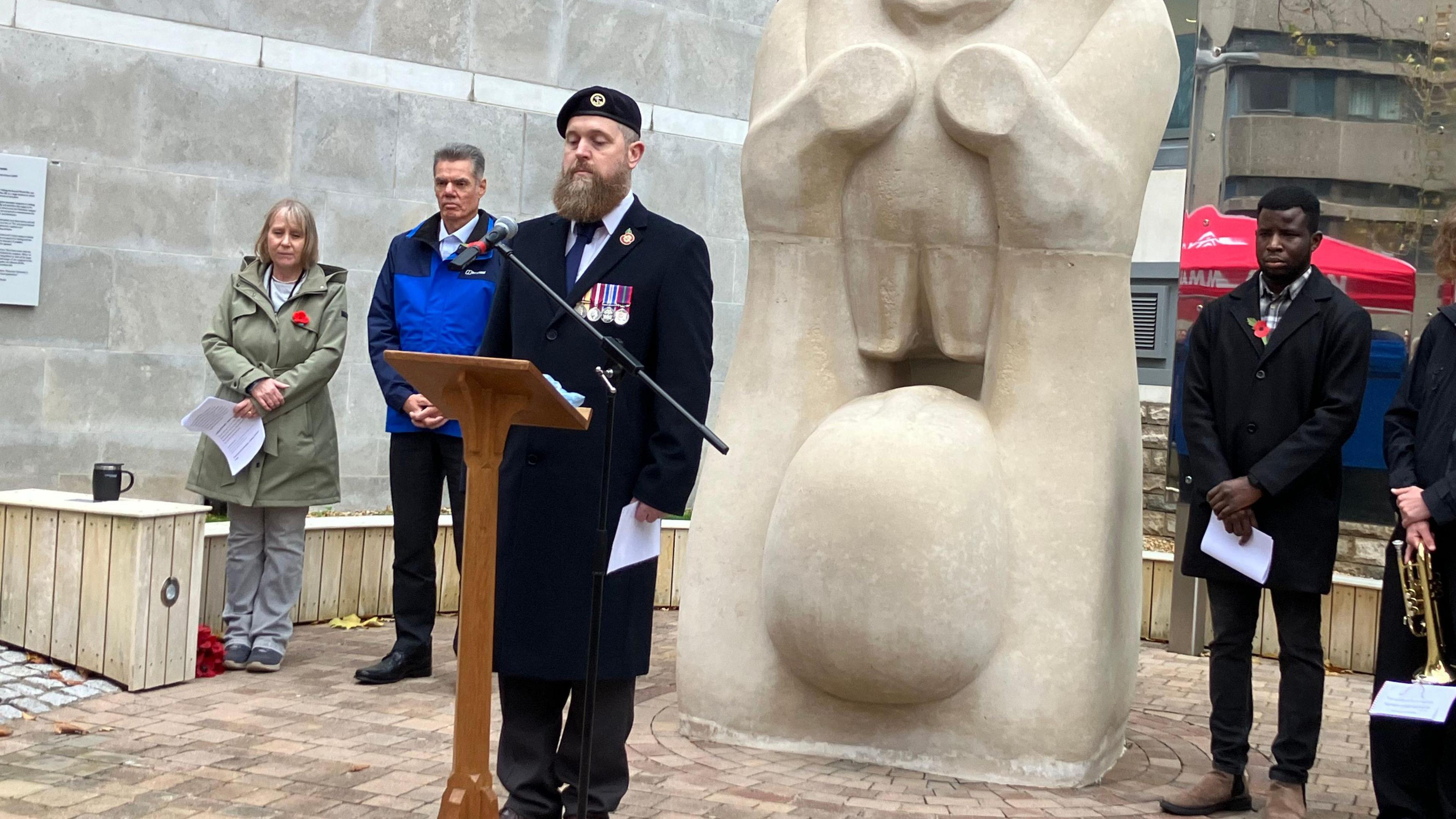 A man wearing uniform is standing in front of a podium with his head bowed for the silence. He is flanked by other people who have their heads bowed.