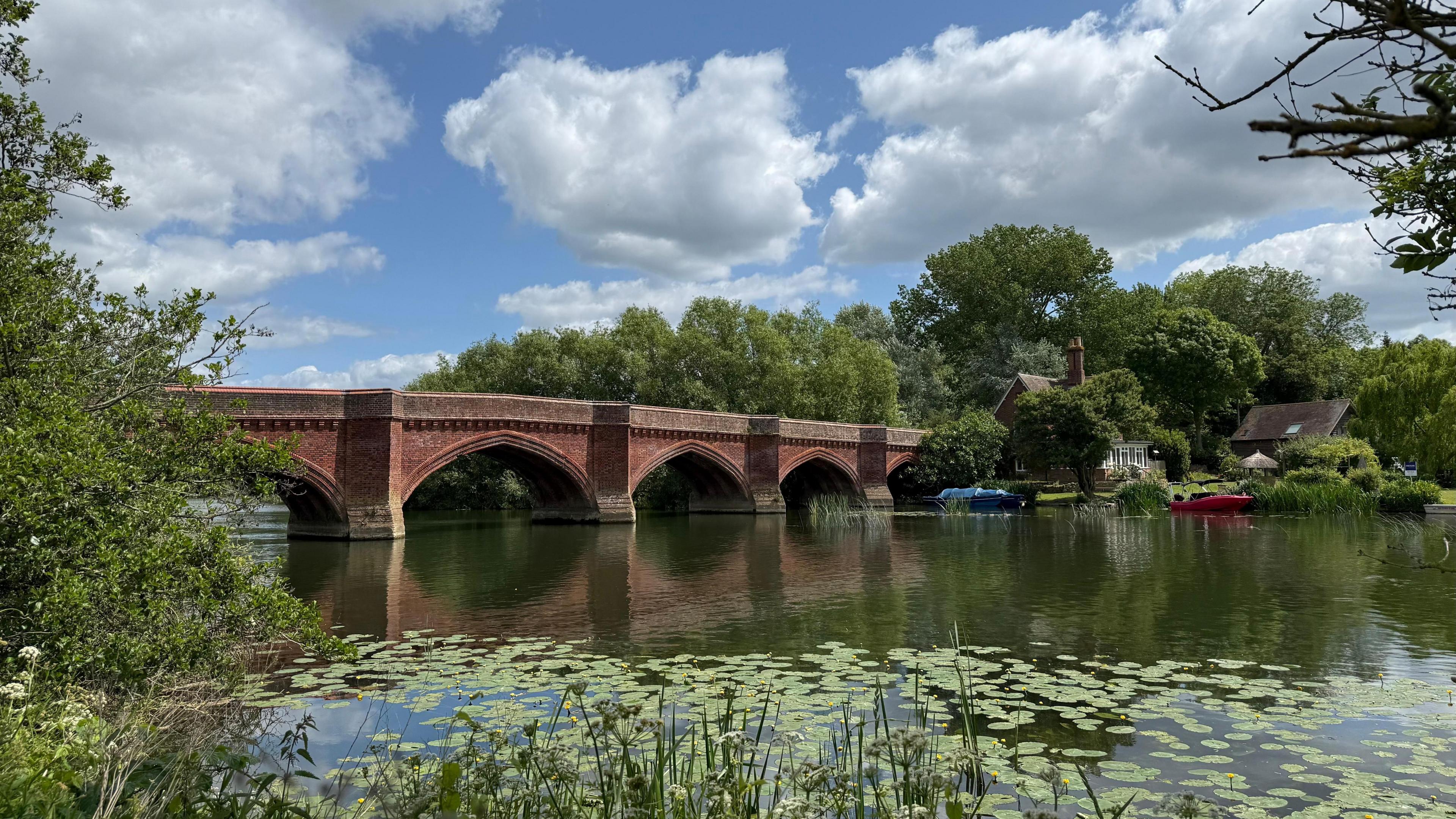 A brick arch bridge over a river on a sunny day. The sky overhead is blue with white clouds. Both banks of the river are lined with trees. There is a building on the far bank with a red and blue boats moored in the water.
