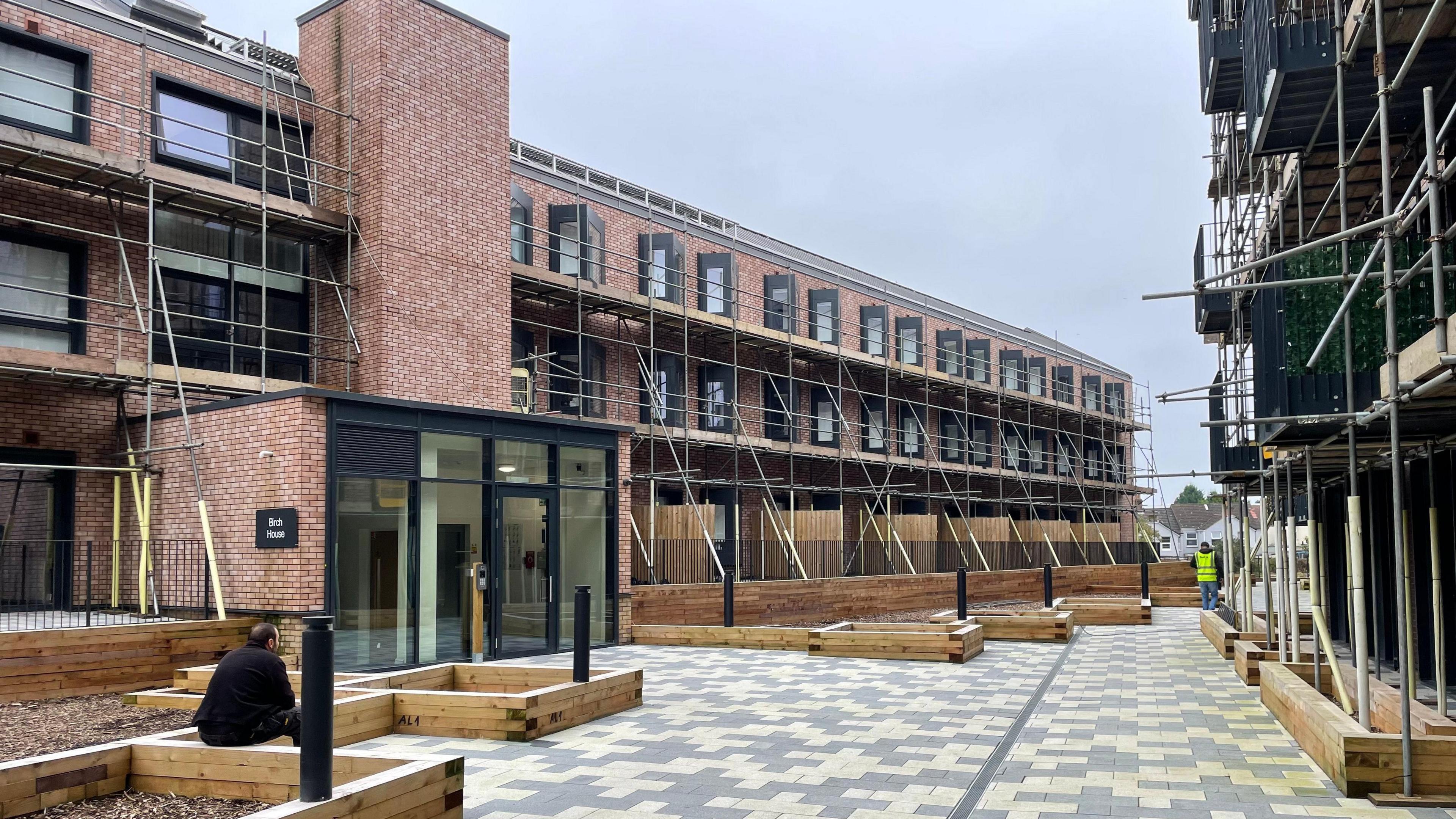 A man sitting on the edge of wooden plant beds which are outside modern three-storey flats which are shrouded in scaffolding. There is a grey and white paved area between two rows of flats.