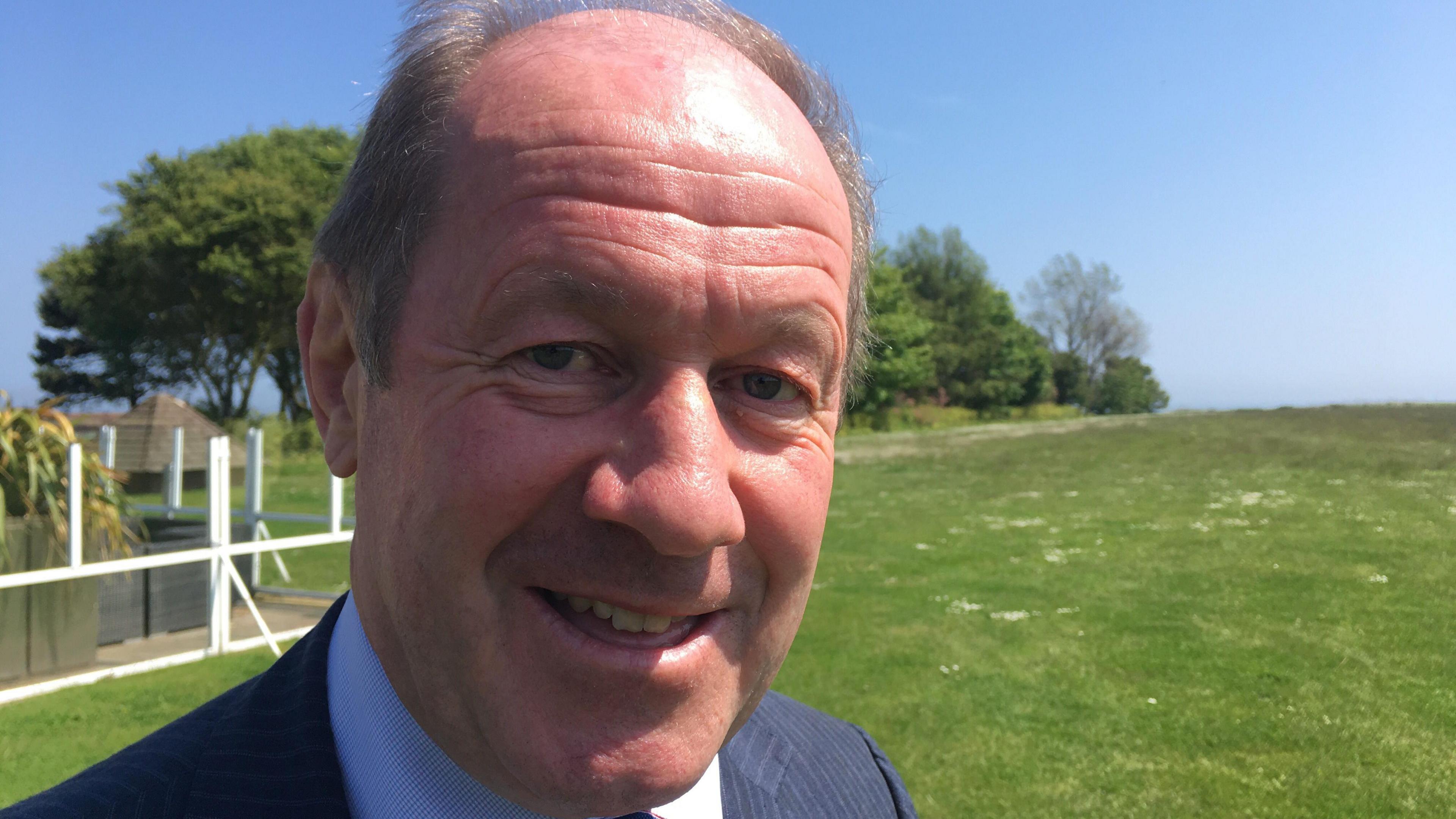 Tim Passmore wearing a blue suit with a red and blue tie, standing on grass with trees in the background.