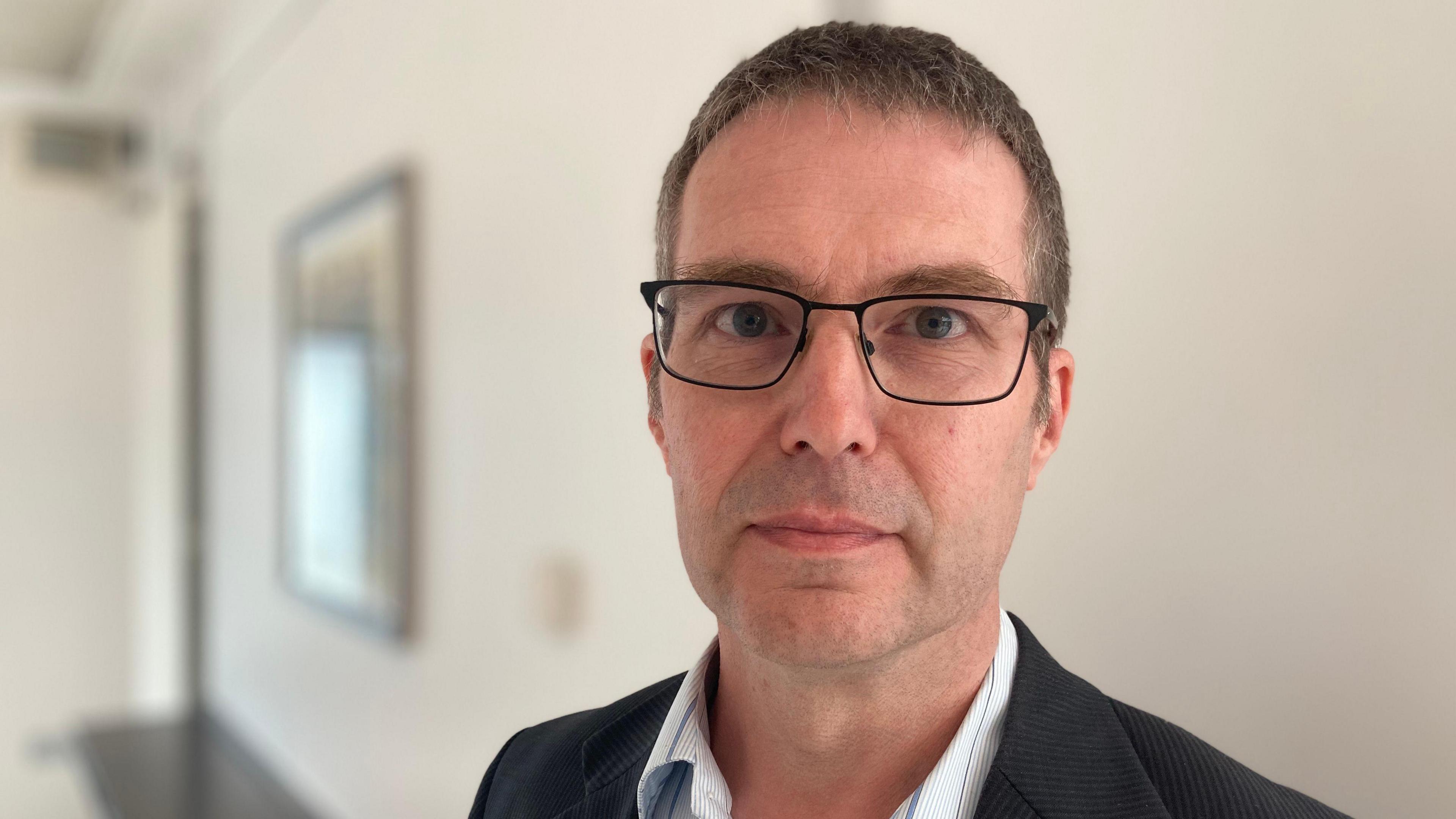 A man is looking into the camera. He is wearing black glasses and a jacket with shirt. He's standing in a large room.