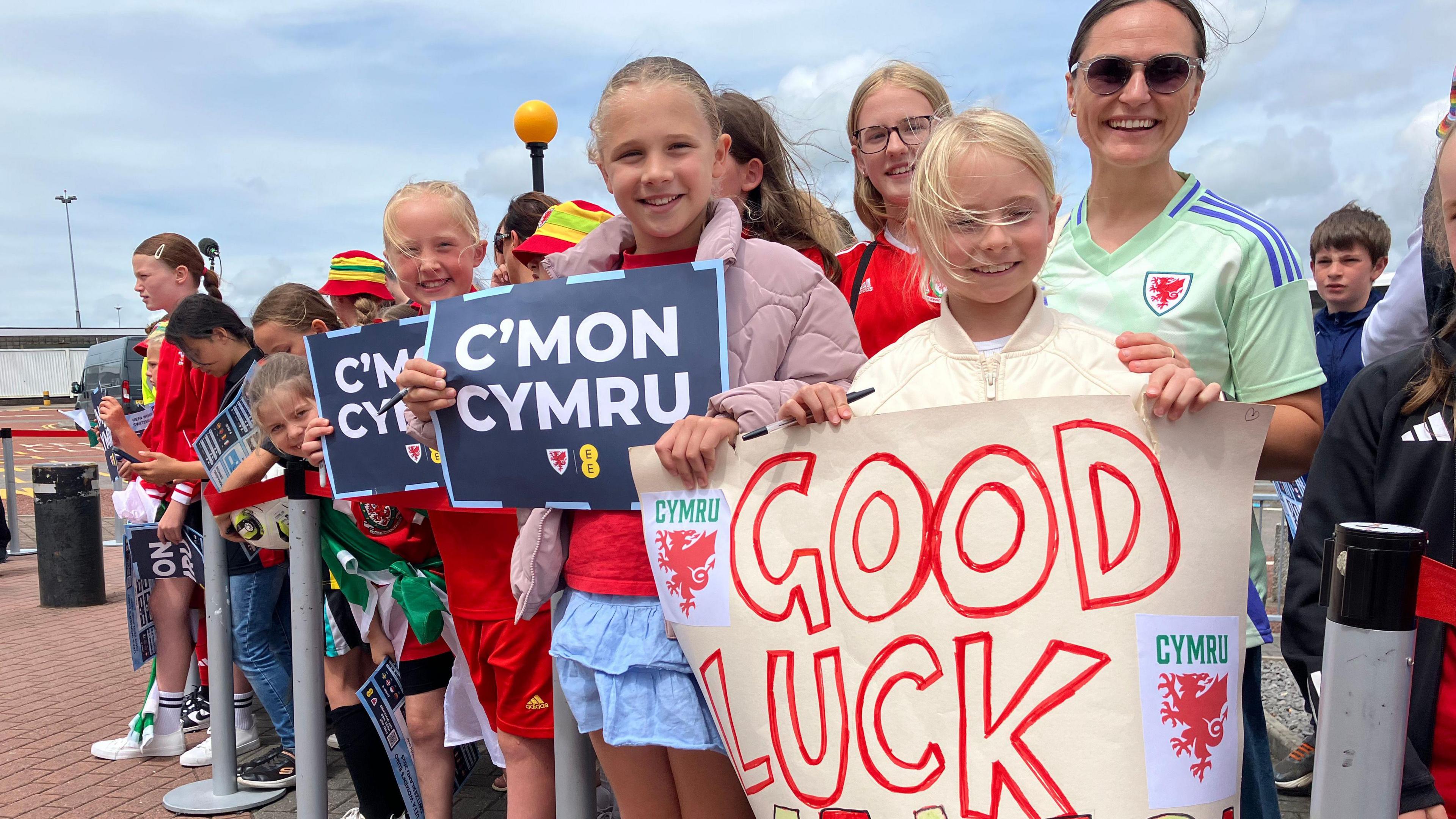 A young girl in the centre in a light brown jacket holds a sign with the words C'mon Cymru. On the right is a girl with blonde hair and a yellow jacket holding a banner with the words Good luck. Behind her is a woman with dark hair and sunglasses wearing a white top.