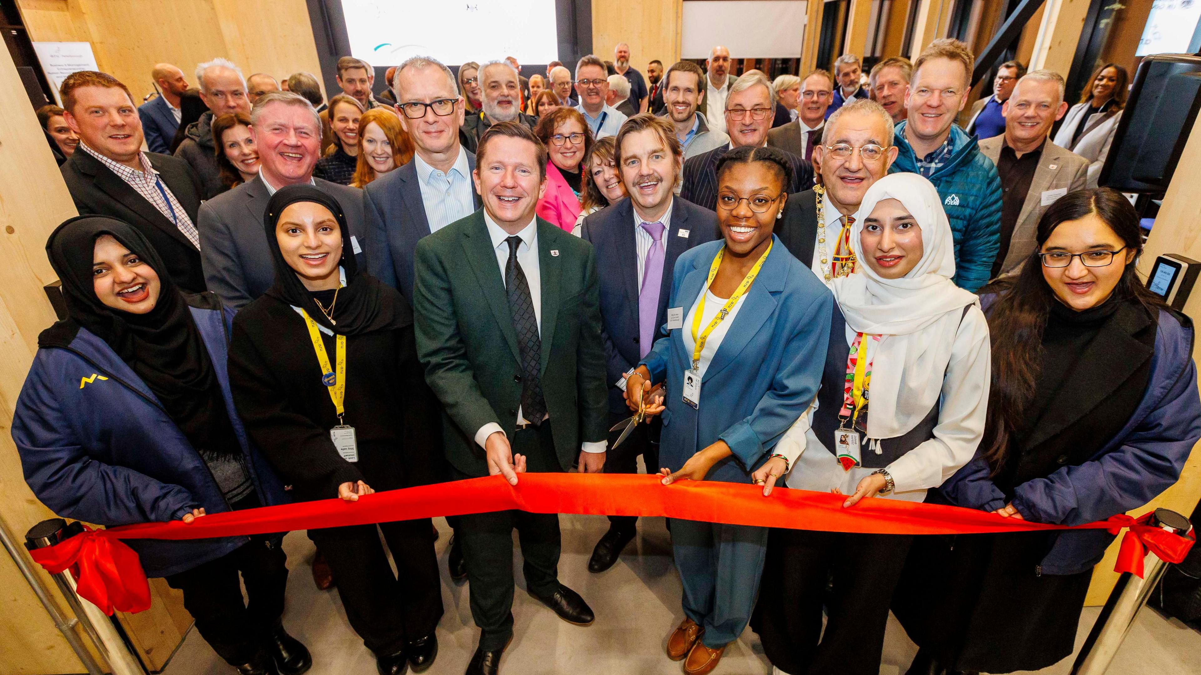 A group of people smile at the camera as a red ribbon is about to be cut to officially open a building.