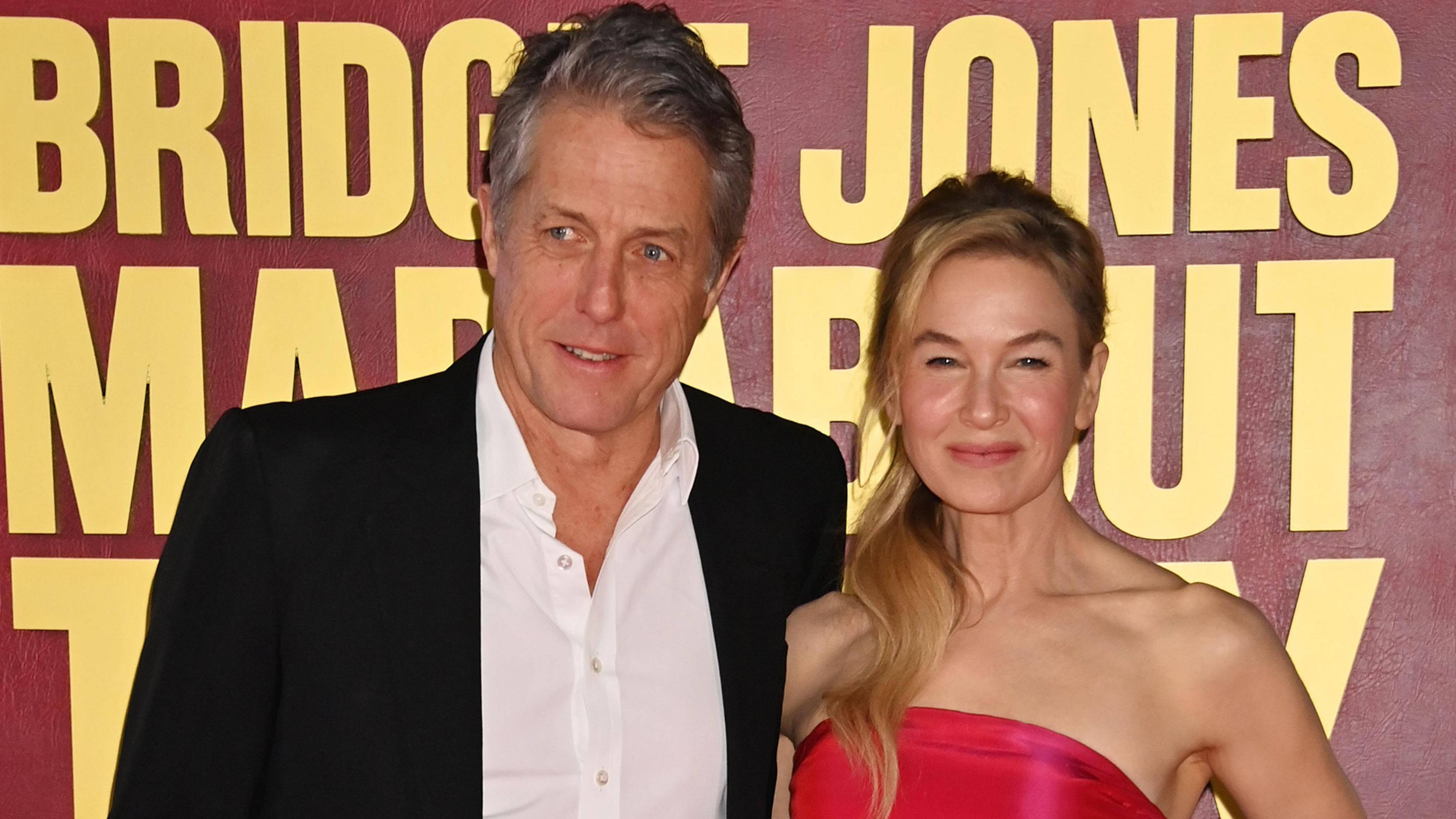 Hugh Grant and Renee Zellweger smile in front of a sign reading Bridget Jones Mad About the Boy