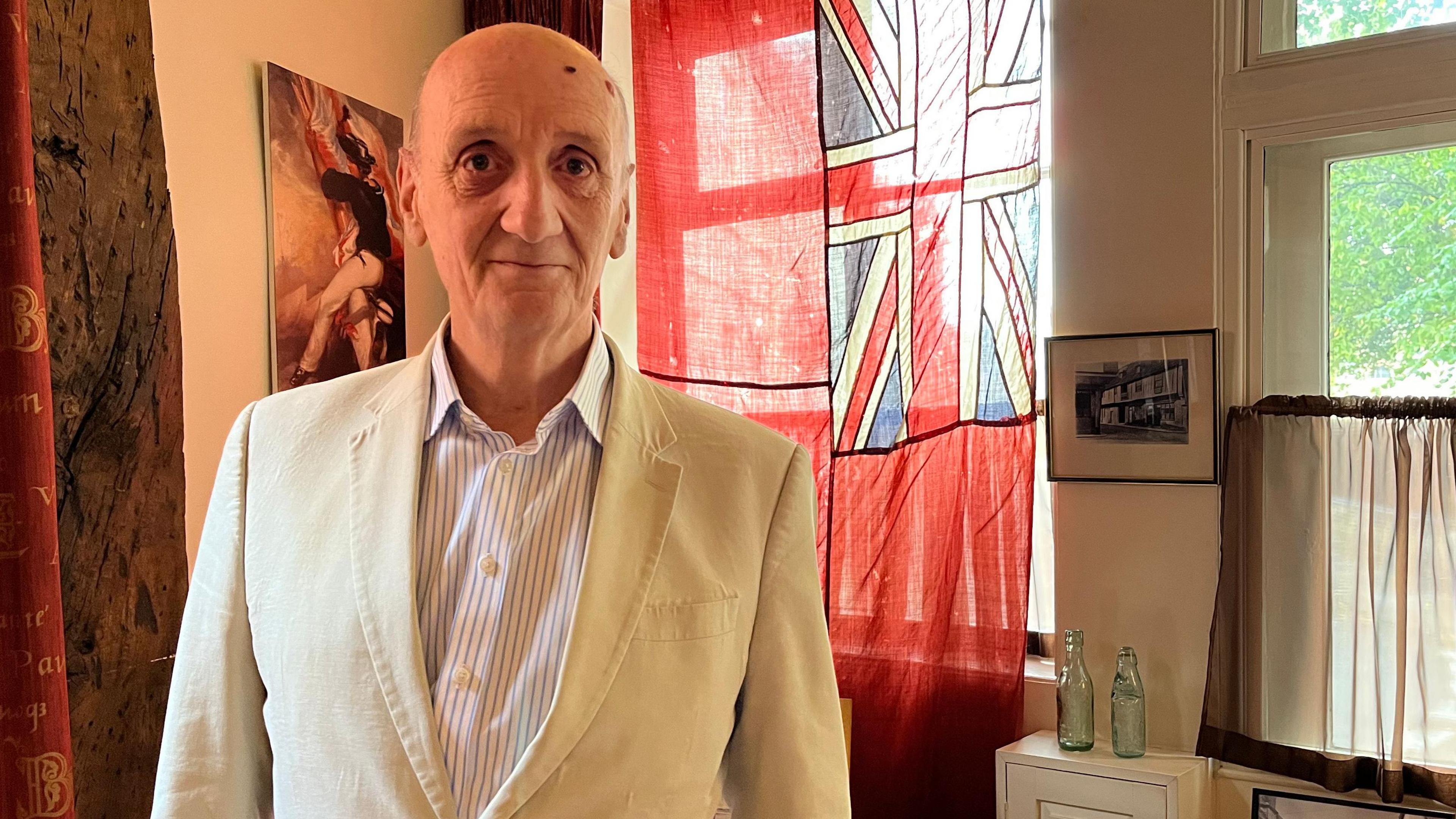 Peter Berthoud stands in front of a red flag with a union flag in its top corner. The room he is in has old wooden beams holding up the roof. Peter is bald, wearing a cream linen suit and white striped shirt.