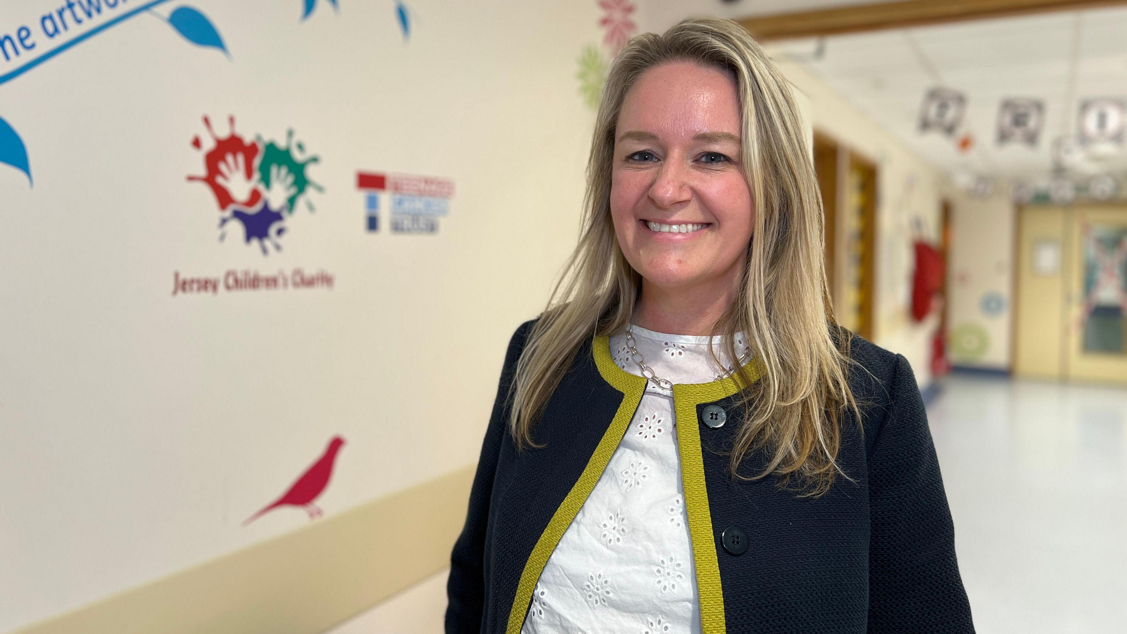 Clair smiles at the camera and she's wearing a dark blue fleece with a yellow trim and a paylsey white top underneath. She's standing in the ward corridoor. Clair has blond hair which comes down to her shoulders.