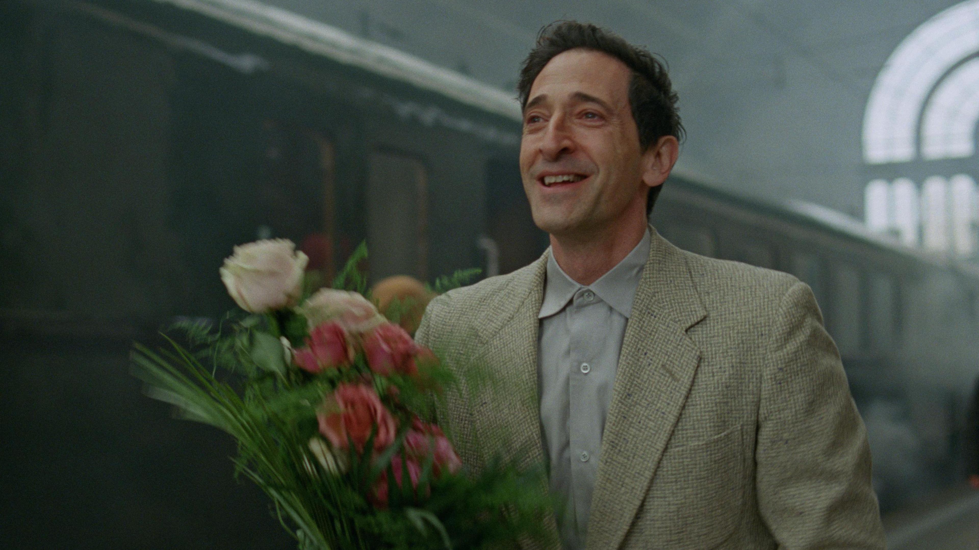 Adrien Brody as László Tóth. He has short dark hair and is wearing a beige suit and grey shirt. He is holding a bunch of pink and white flowers and smiling as he walks along a train platform with a train behind him.