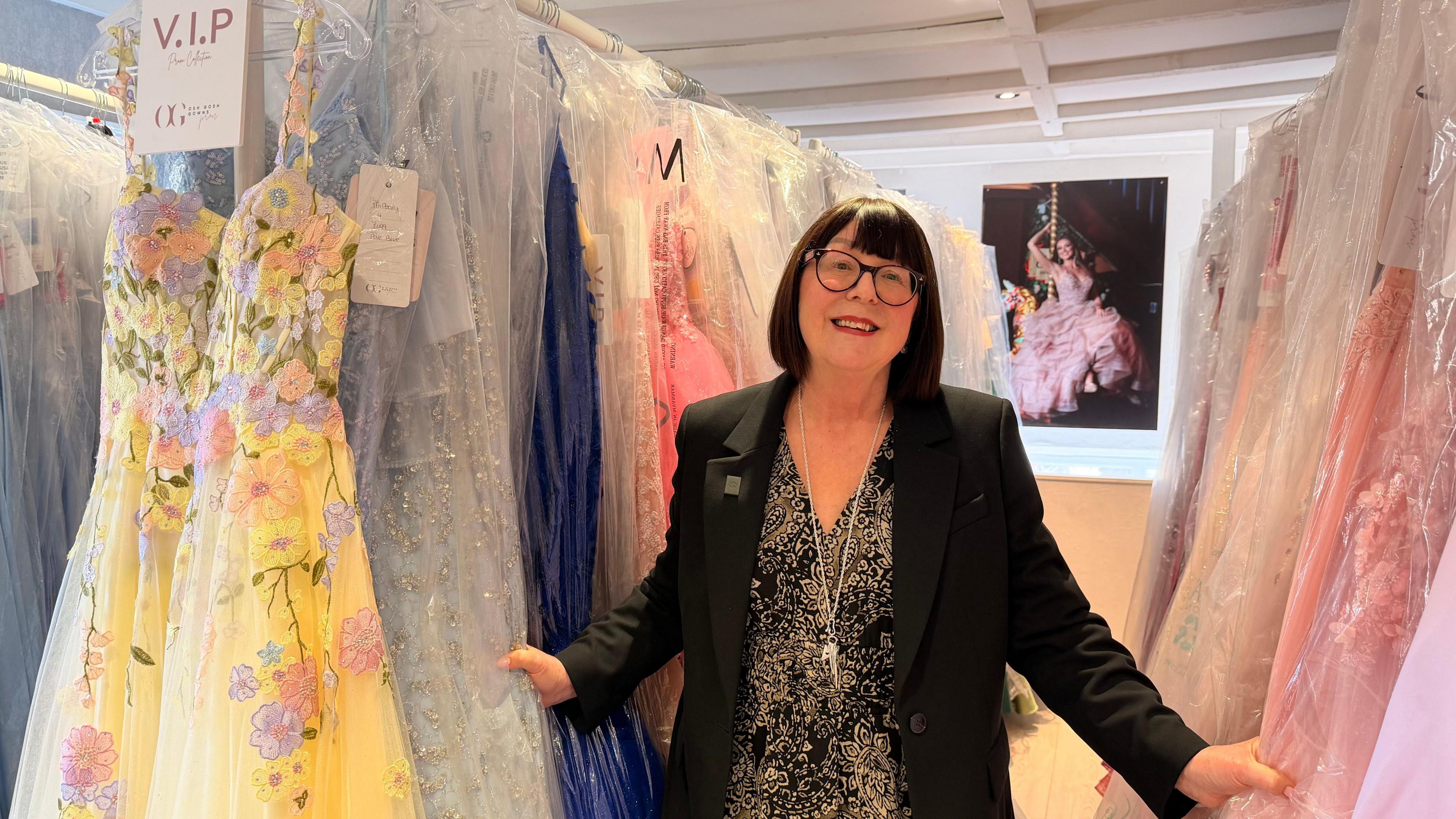 A woman smiles as she stands between two racks of prom dresses in plastic wrappers, with pink, yellow and blue colours on show. She has a short black bob and a fringe and wears a black and gold floral dress, black blazer and round-rimmed glasses. In the background, a print of a woman on a swing in an elaborate pink gown can be seen.