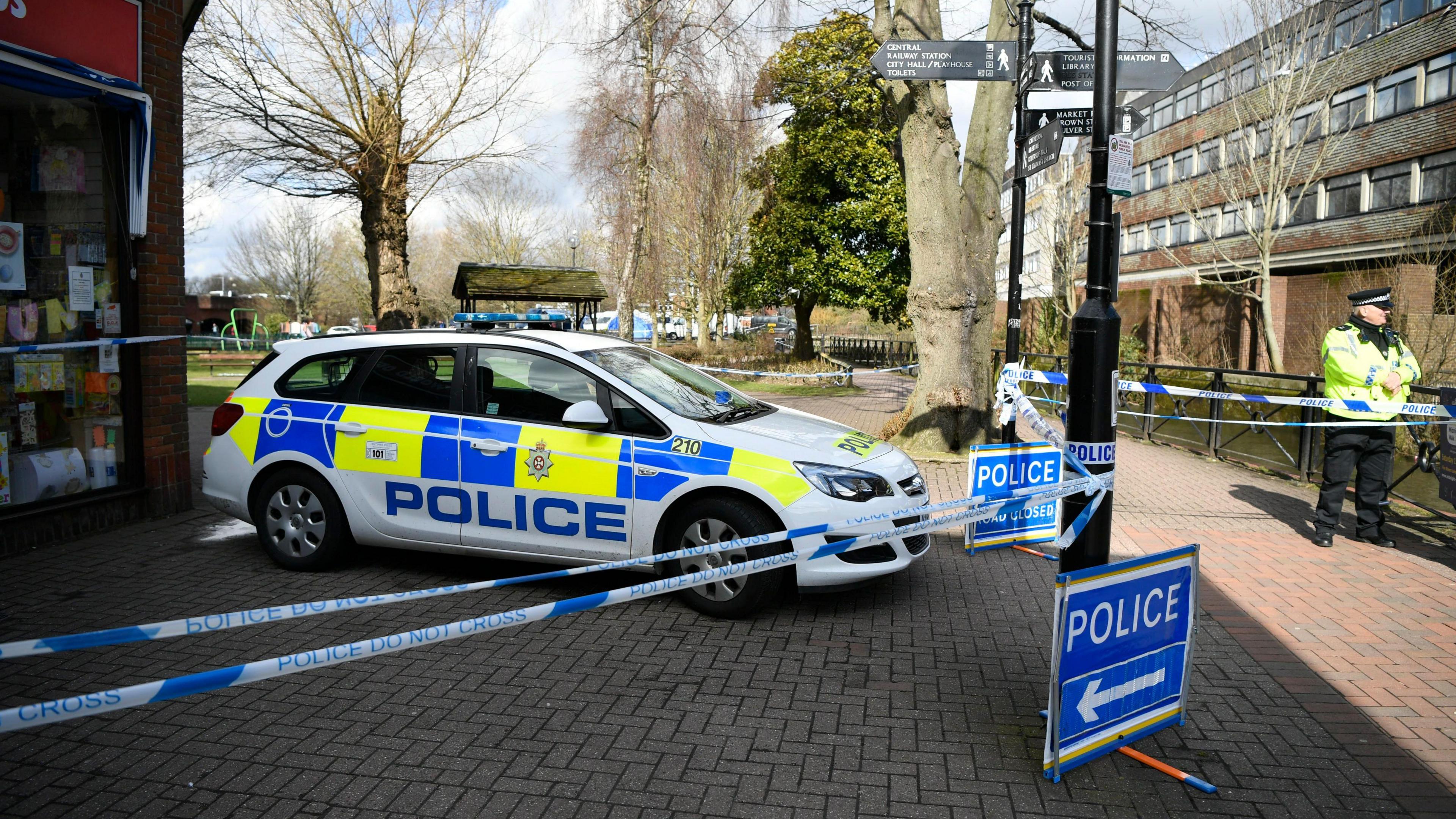 A police car behind a police cordon in Salisbury, parked on a brick paved road with parts of buildings visible on either side and treess and a park in the background. A police officers in a yellow high-vis jacket an dflat cap stands to the left hand side of the photo behind the police cordon tape