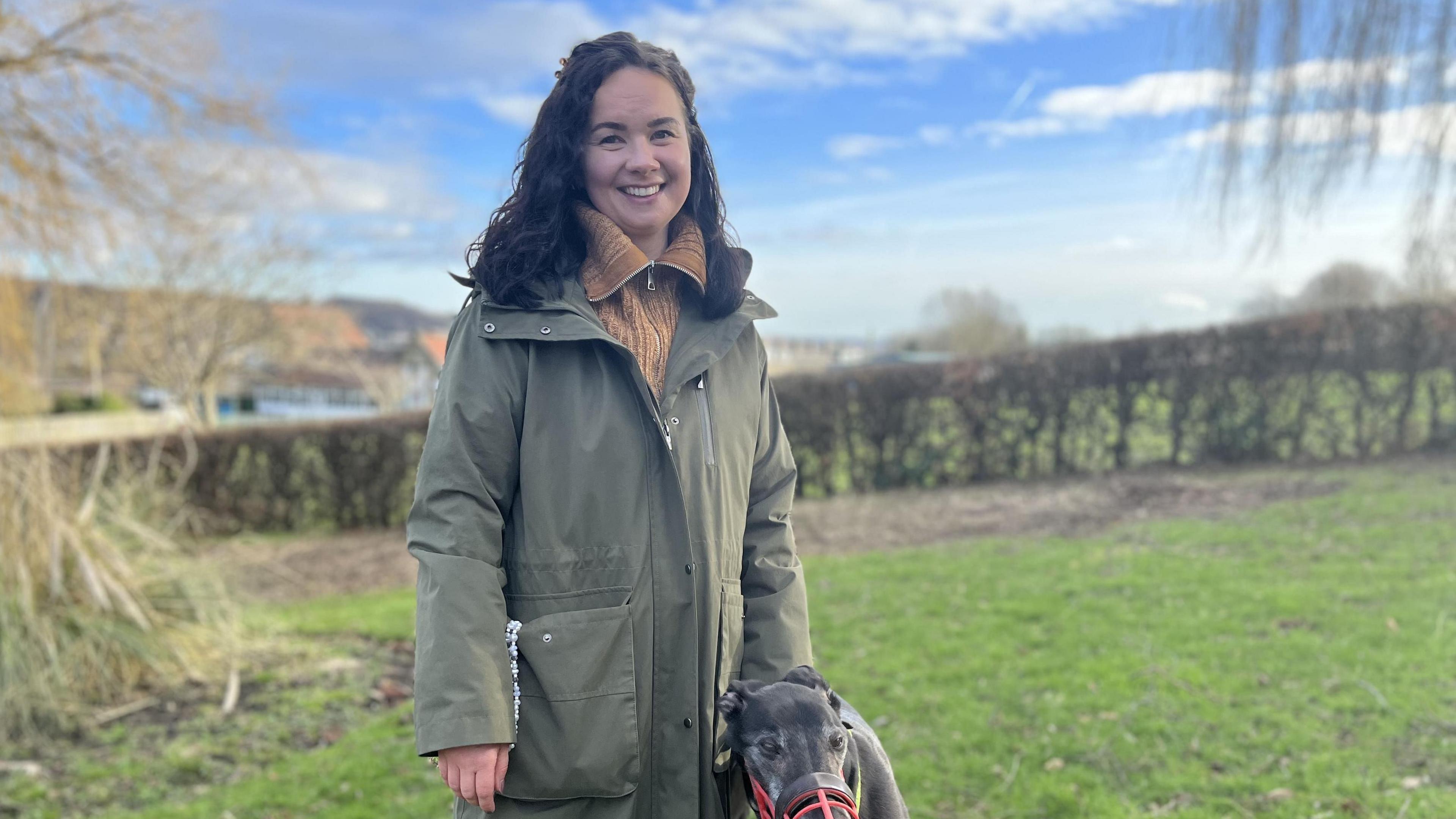 A young woman in a long green coat stands smiling in a field with her pet dog, a grey whippet.