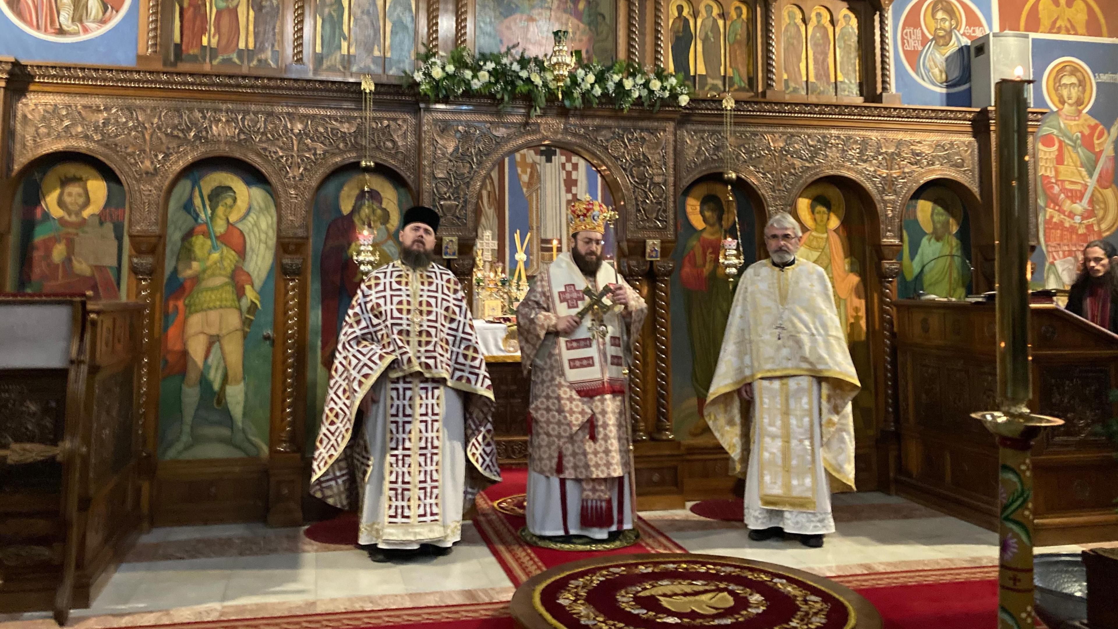 Inside the church. Three church leaders dressed in religious robes. The man in the middle holds a cross and behind them are colourful paintings of figures and patterned arches, relics and decorations. 