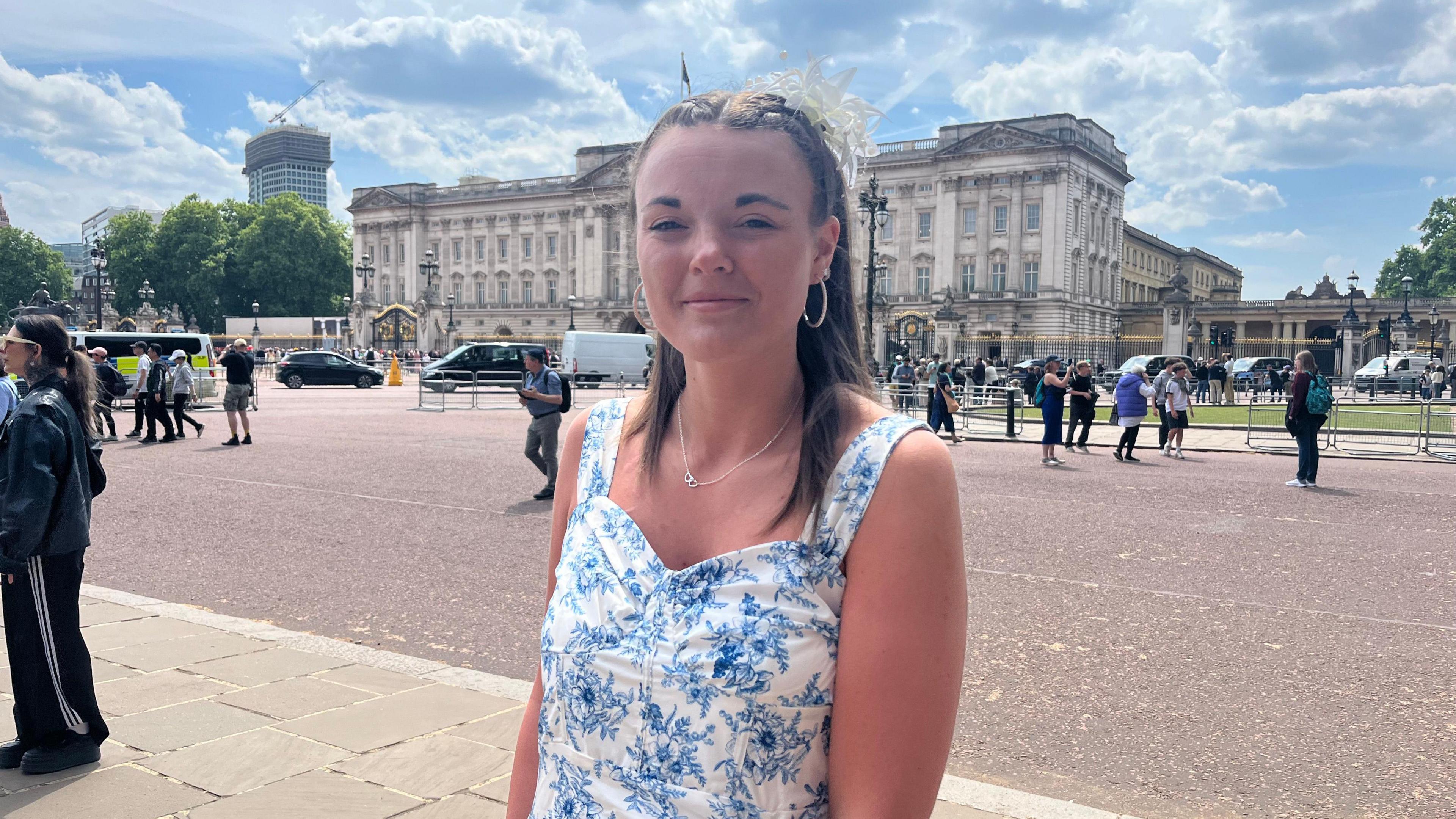 A woman with long brown hair wearing a blue and white patterned dress smiling at the camera. She is stood in front of Buckingham Palace. People can be seen gathered or walking in the background. The sky is sunny with white clouds.
