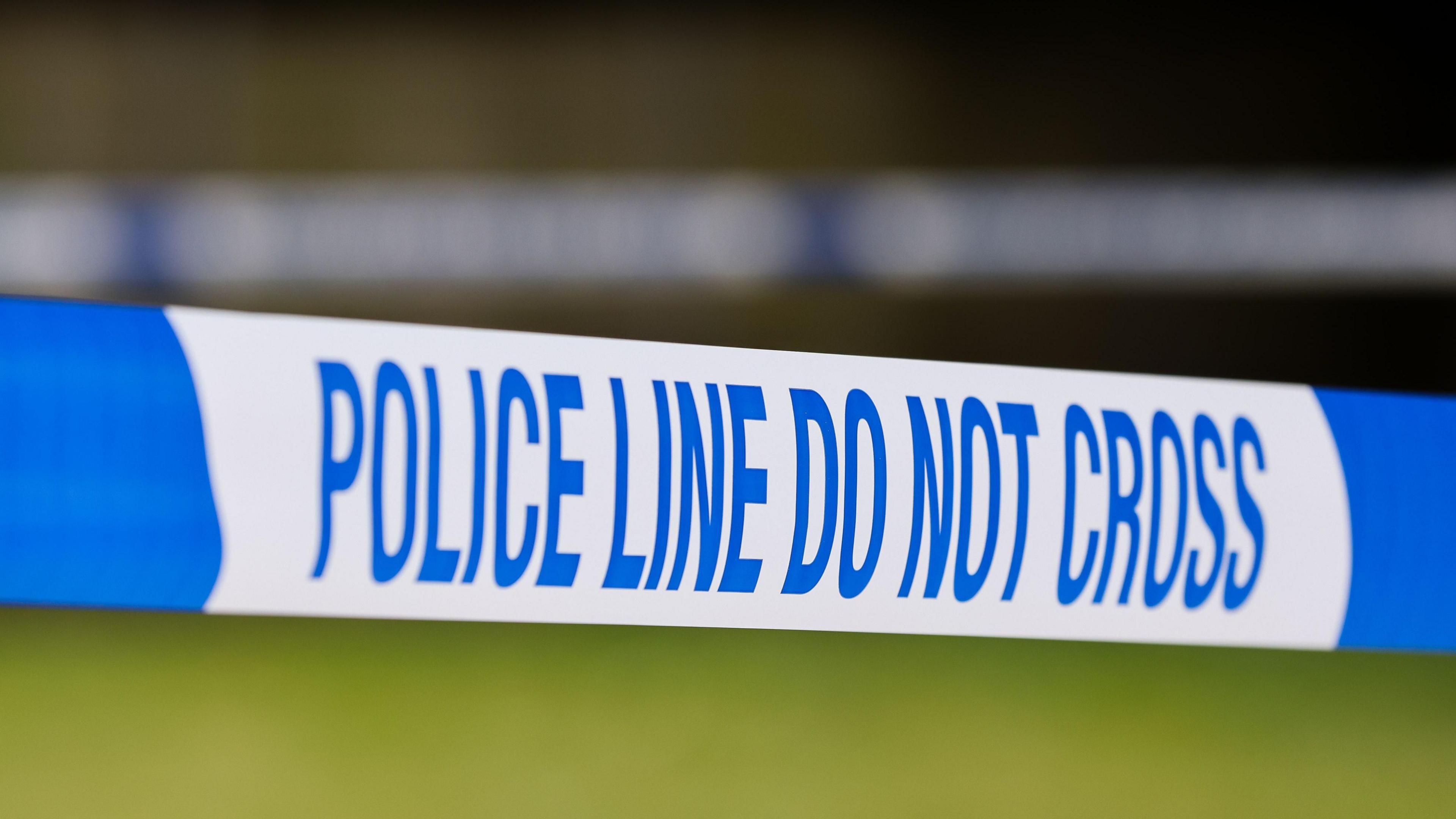 Taut blue-and-white police tape marked with the words: "Police line do not cross", against a blurred green background.