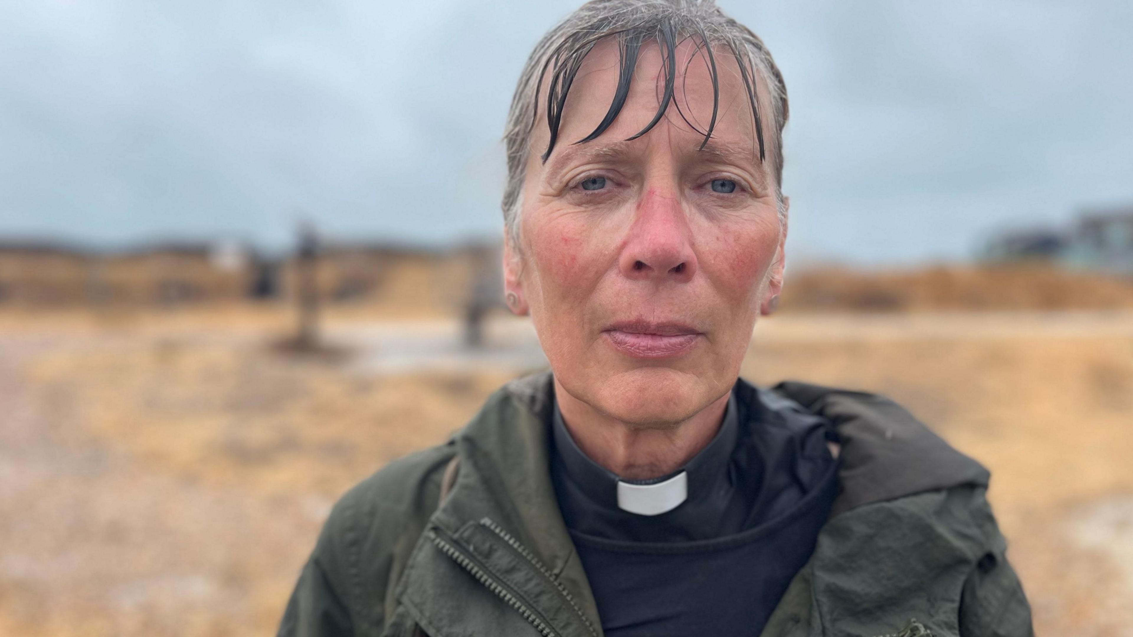 The Rev Jane Bartlett stands on a rainy beach near Shoreham seafront wearing a dark coat and clerical collar. Pebbles, beach vegetation and a grey sky can be seen in the background.