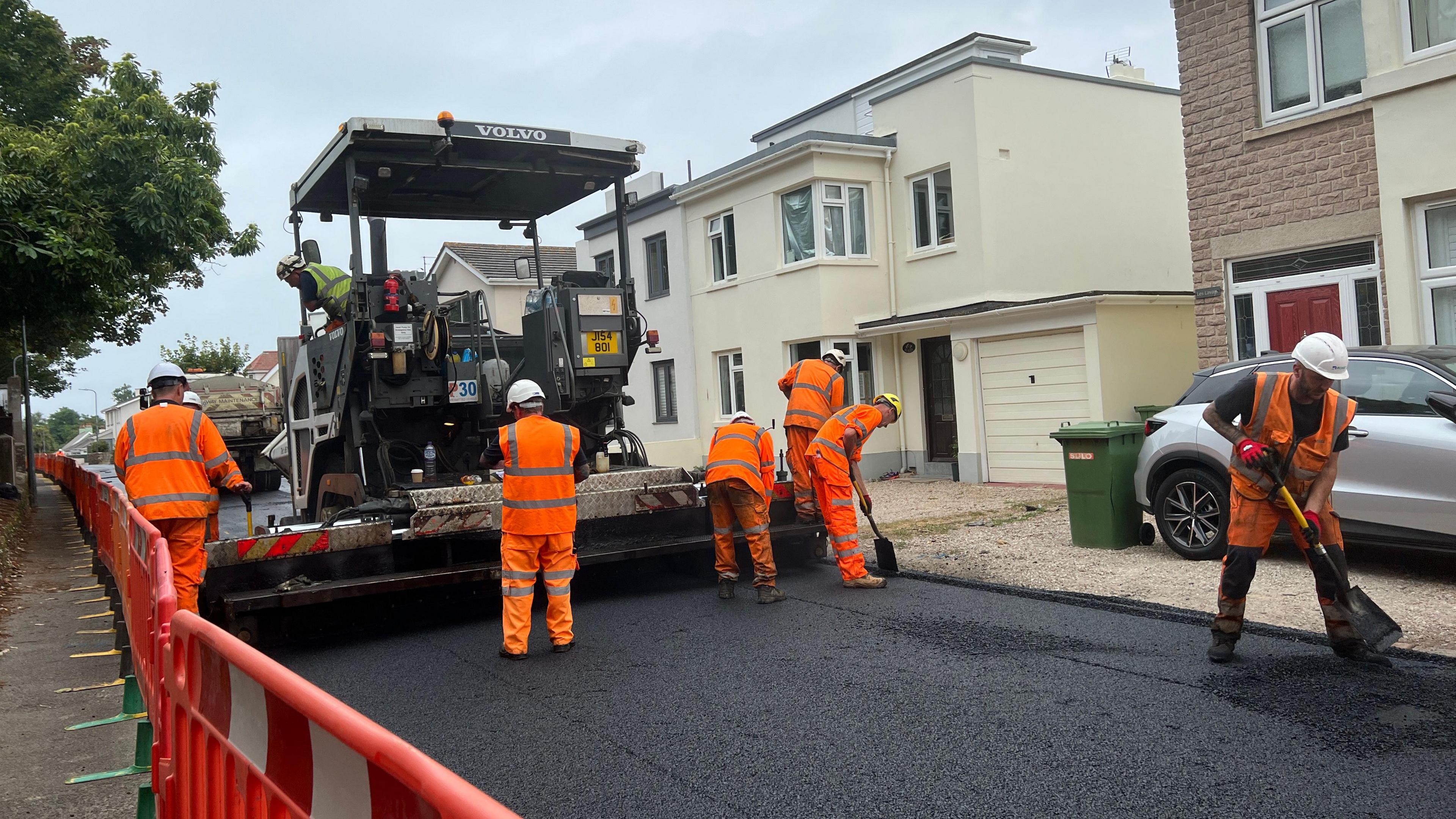 Six men wearing orange high vis workwear tarmacking the road with a big machine. Temporary railings on the left and houses on the right. Grey skies.
