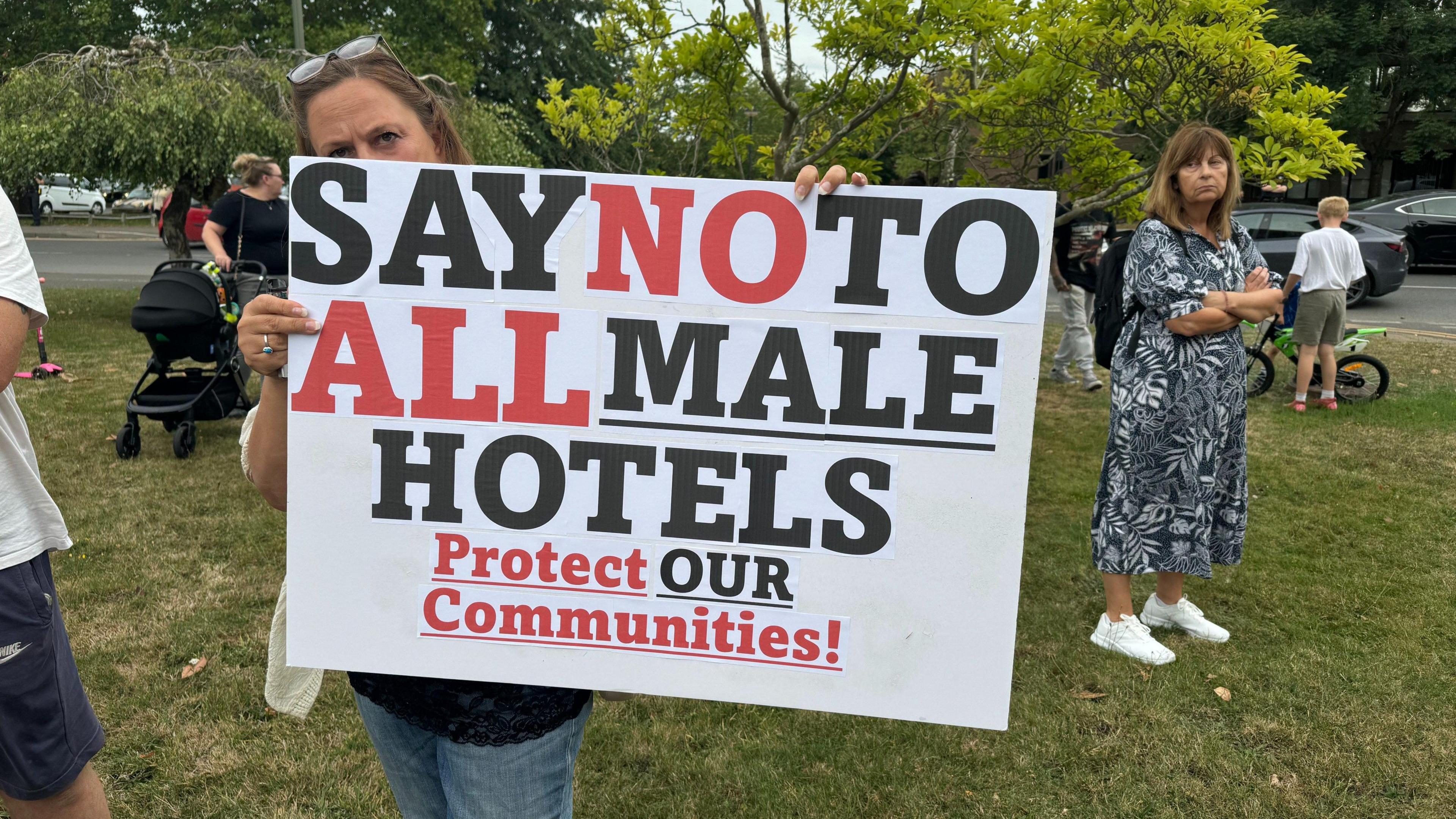 A female protestor holds up a sign saying "Say no to all male hotels. Protect our communities. Several other protestors stand behind her.