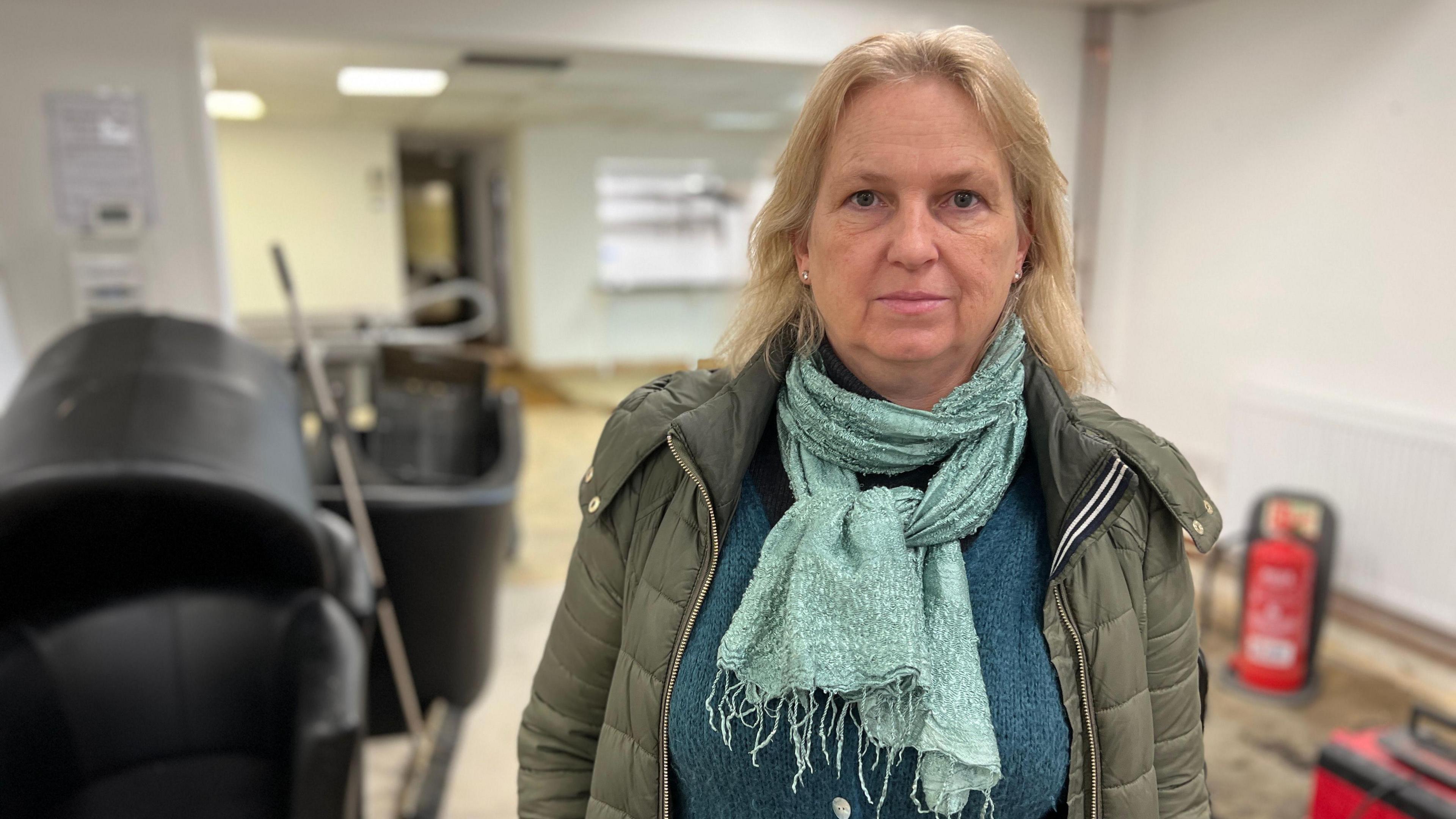 Jo Kitching stands in the hall of The Citadel. She has short blonde hair and is wearing a puffer jacket, turquoise jumper and a turquoise scarf. 