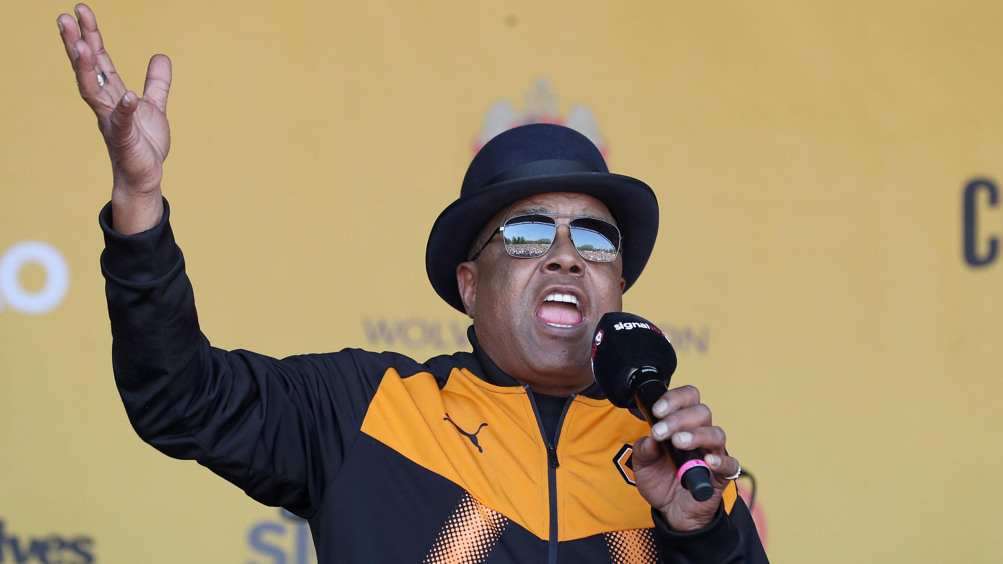 Tito Jackson in a black bowler hat wearing sunglasses and a mustard yellow and black Wolverhampton Wanderers hoodie, as he sings into a mic