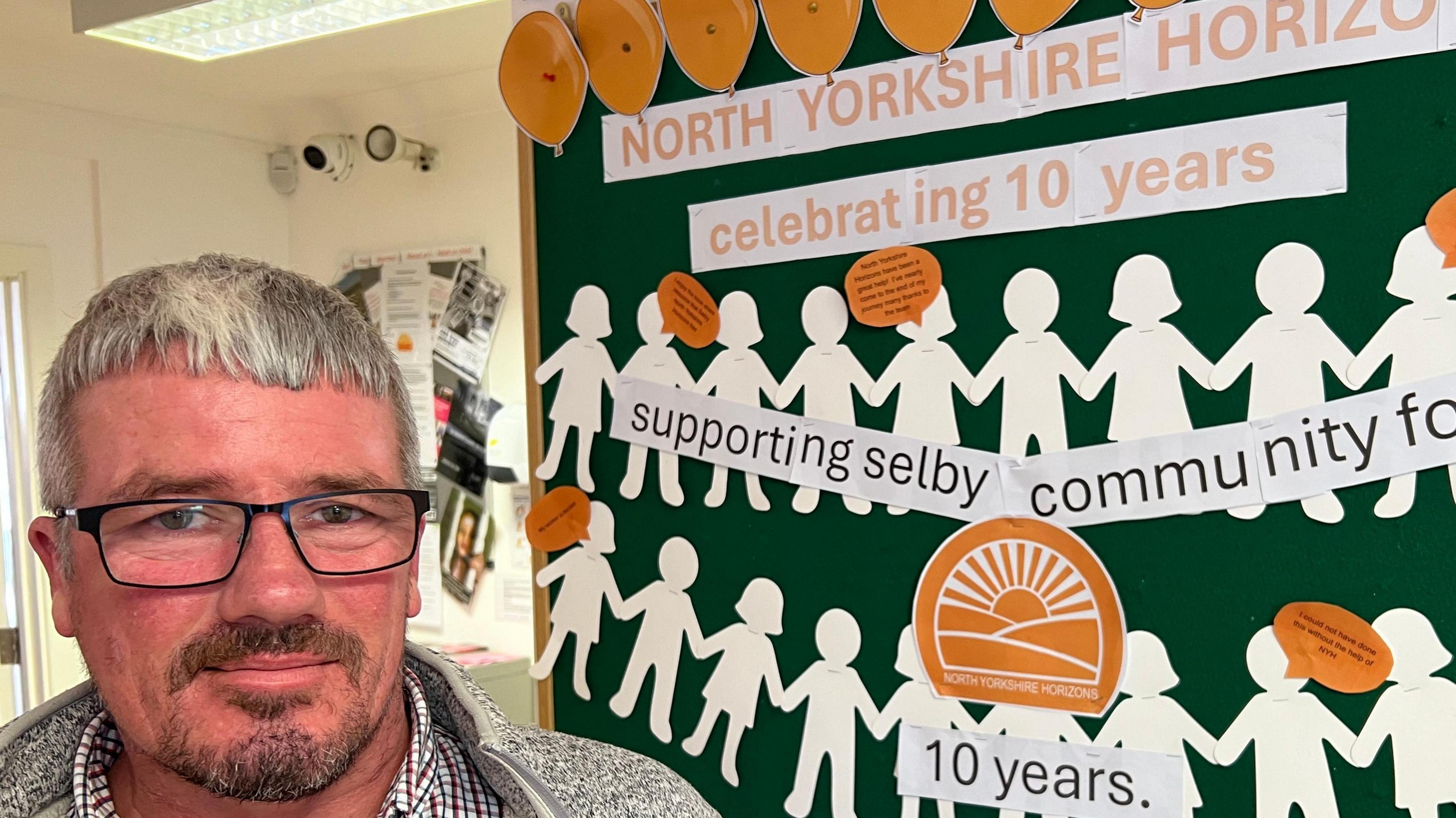 Mark has grey hair, is wearing spectacles, and looks at the camera next to a poster marking the charity's tenth anniversary.