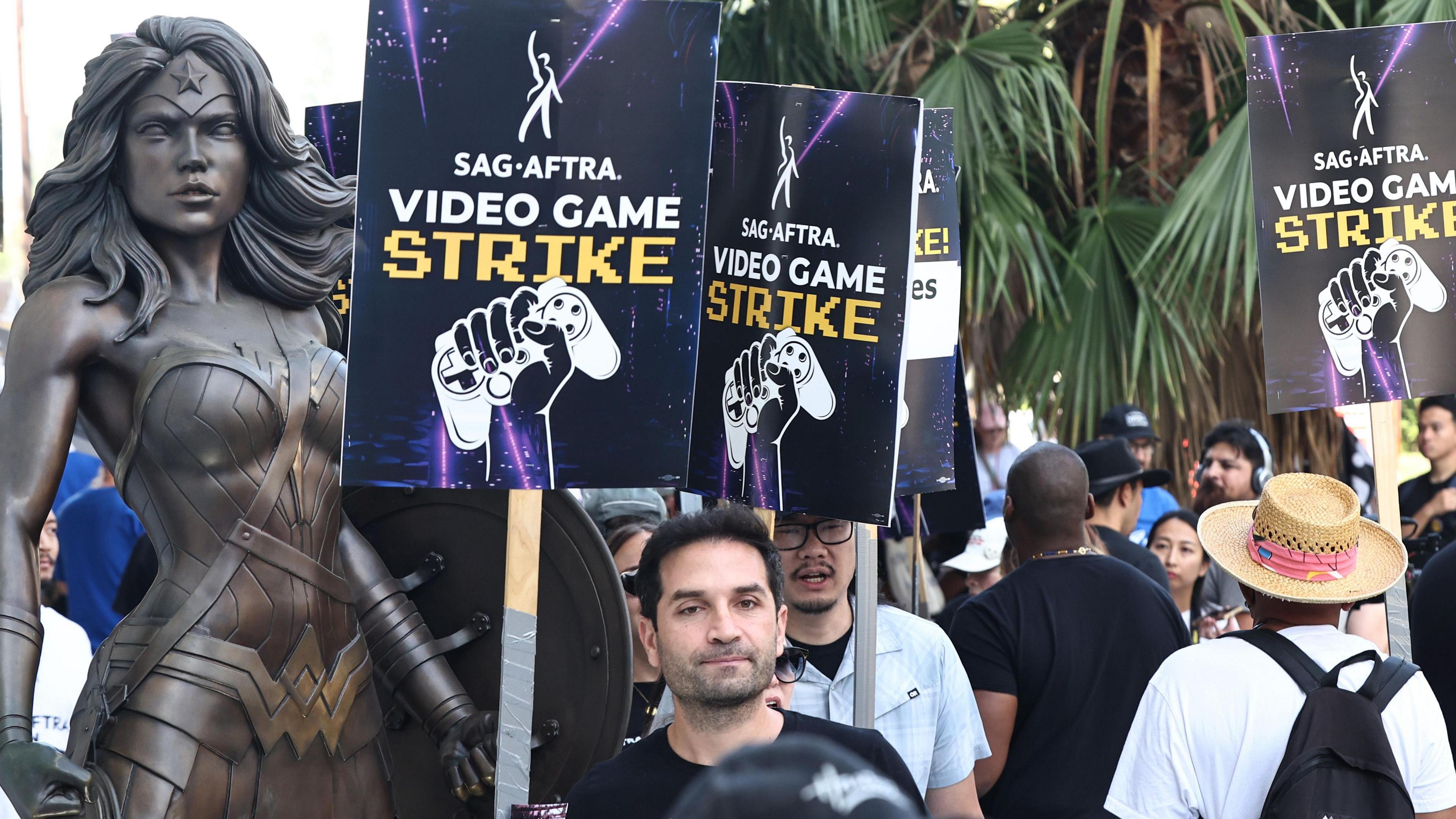 Members of SAG-AFTRA striking at WB Studios. They're holding placards saying "SAG-AFTRA Video game strike" with a hand holding a games controller - in front of a wonderwoman statue. 