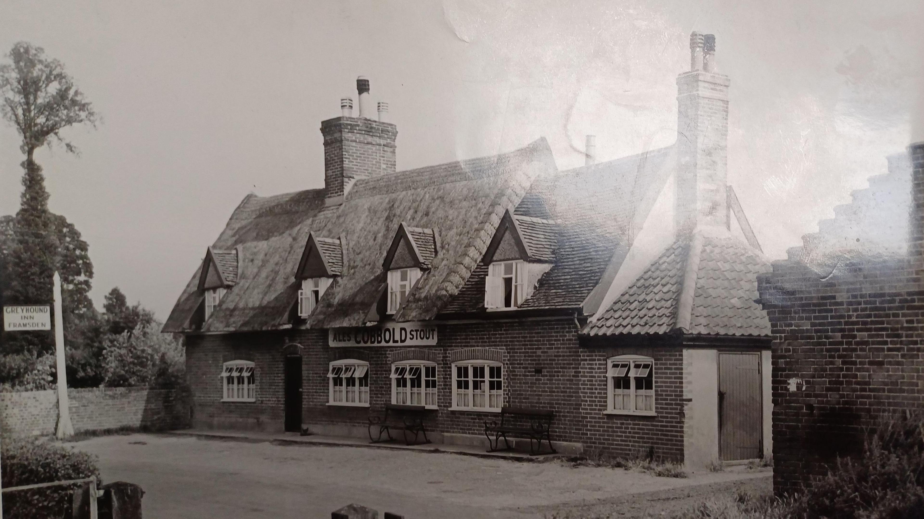 Picture shows a black and white image of the pub with a thatched roof, a sign advertising Cobbold ales and stout as well as a hanging sign saying Greyhound Inn, Framsden.