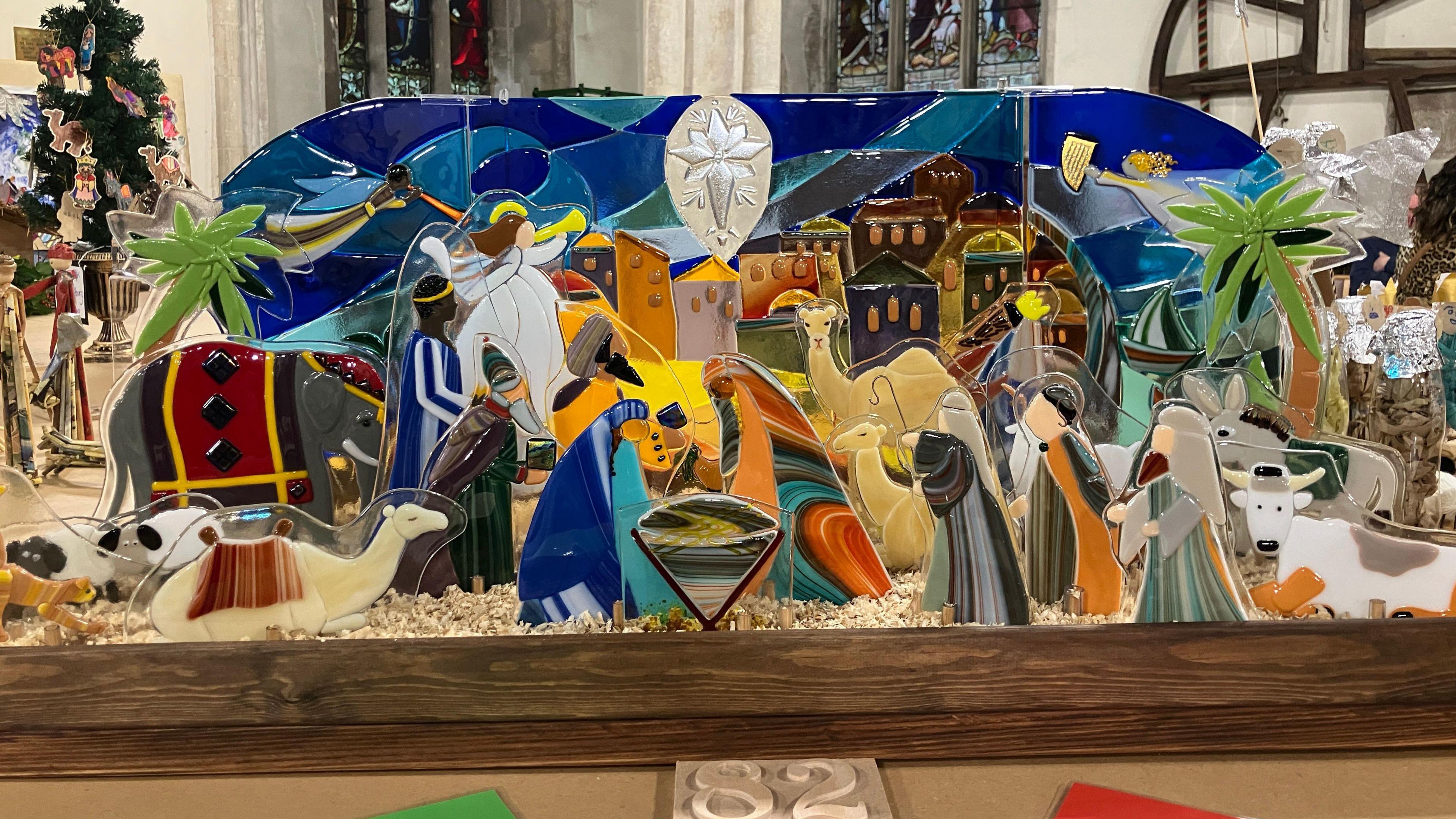 A nativity scene made out of stained glass. Mary and Joseph are in the middle, looking over baby Jesus. Kings, camels, elephants and angels can also be seen in the nativity. 