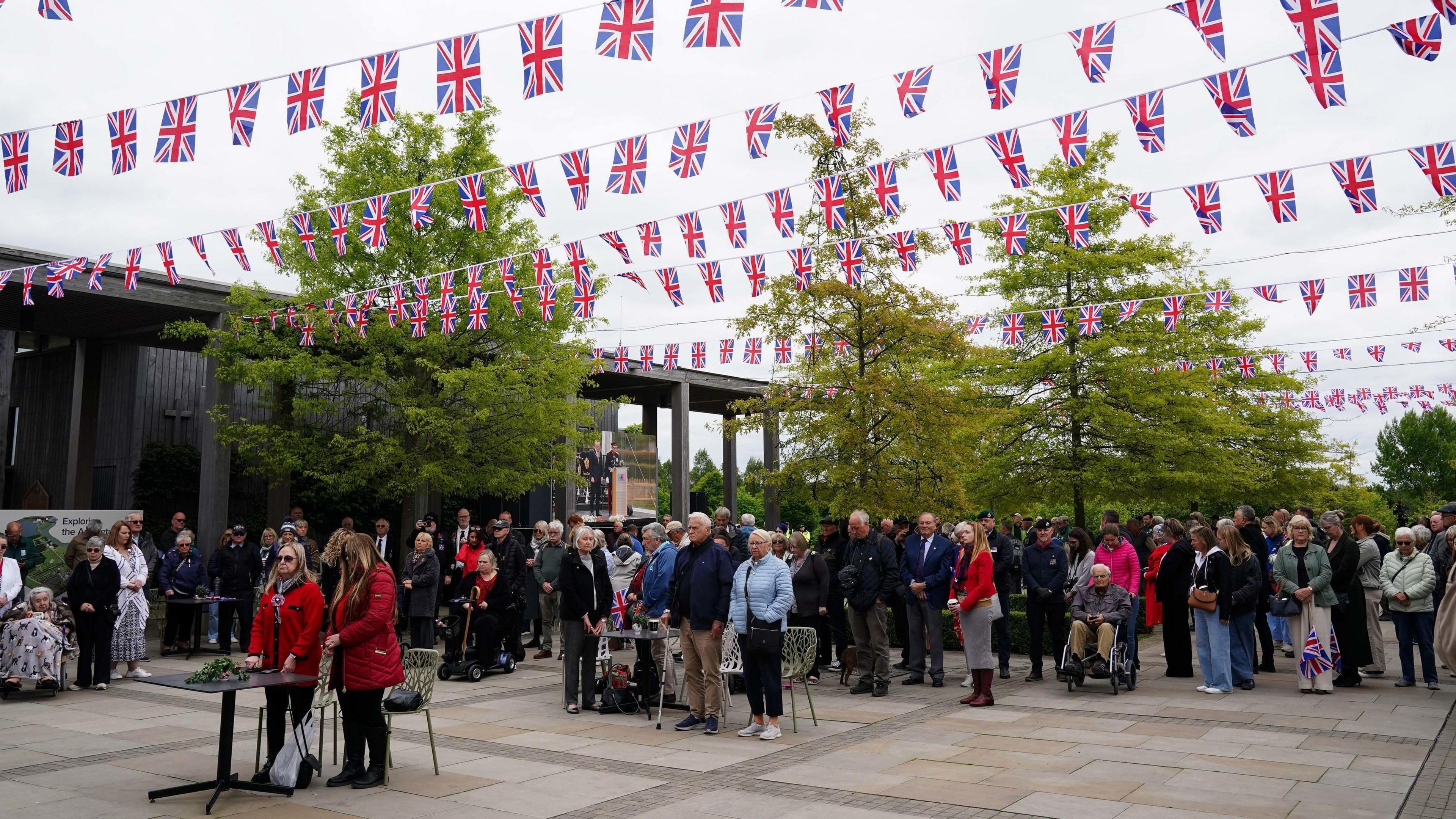 People gathered in groups on a square with union jack bunting above them as they stand silent to mark VE Day.