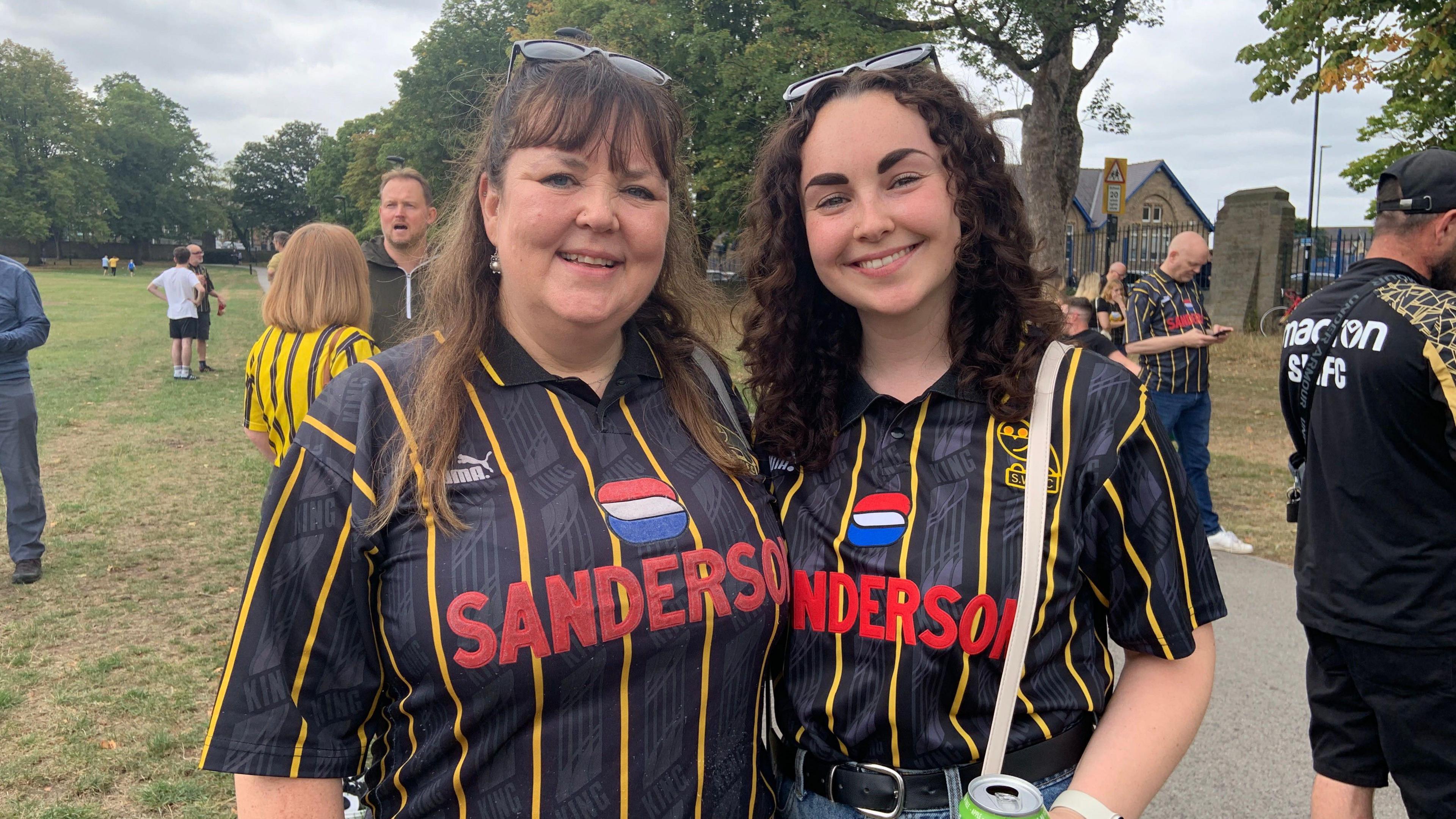 A woman with long straight brown hair is stood next to another woman with dark brown curly hair. Both are wearing black and yellow striped Sheffield Wednesday tops and are stood in a park similing