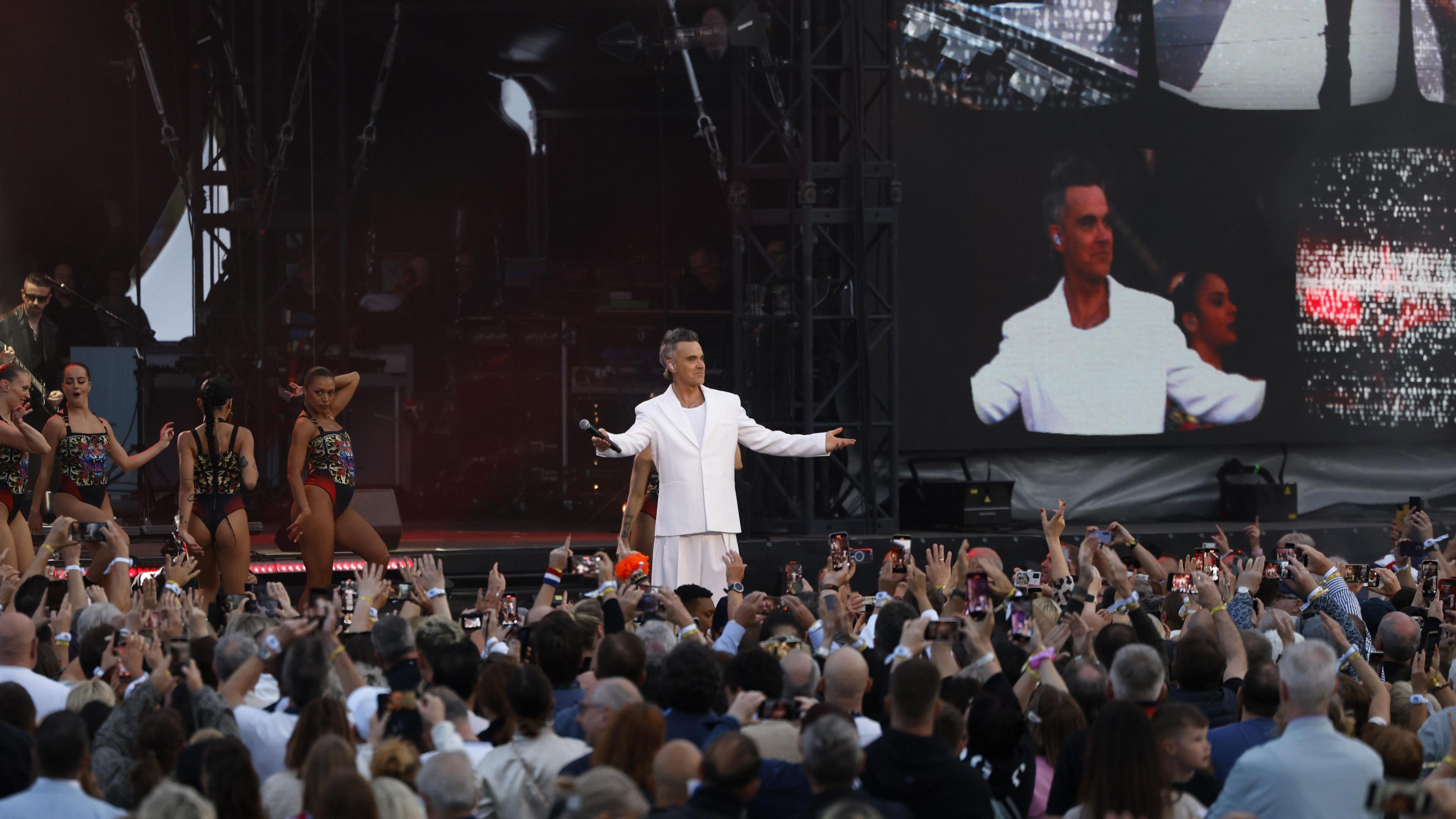 Robbie Williams is dressed in an all-white suit on a stage in front of thousands. There are backing dancers dressed in sparkly costumes behind him.