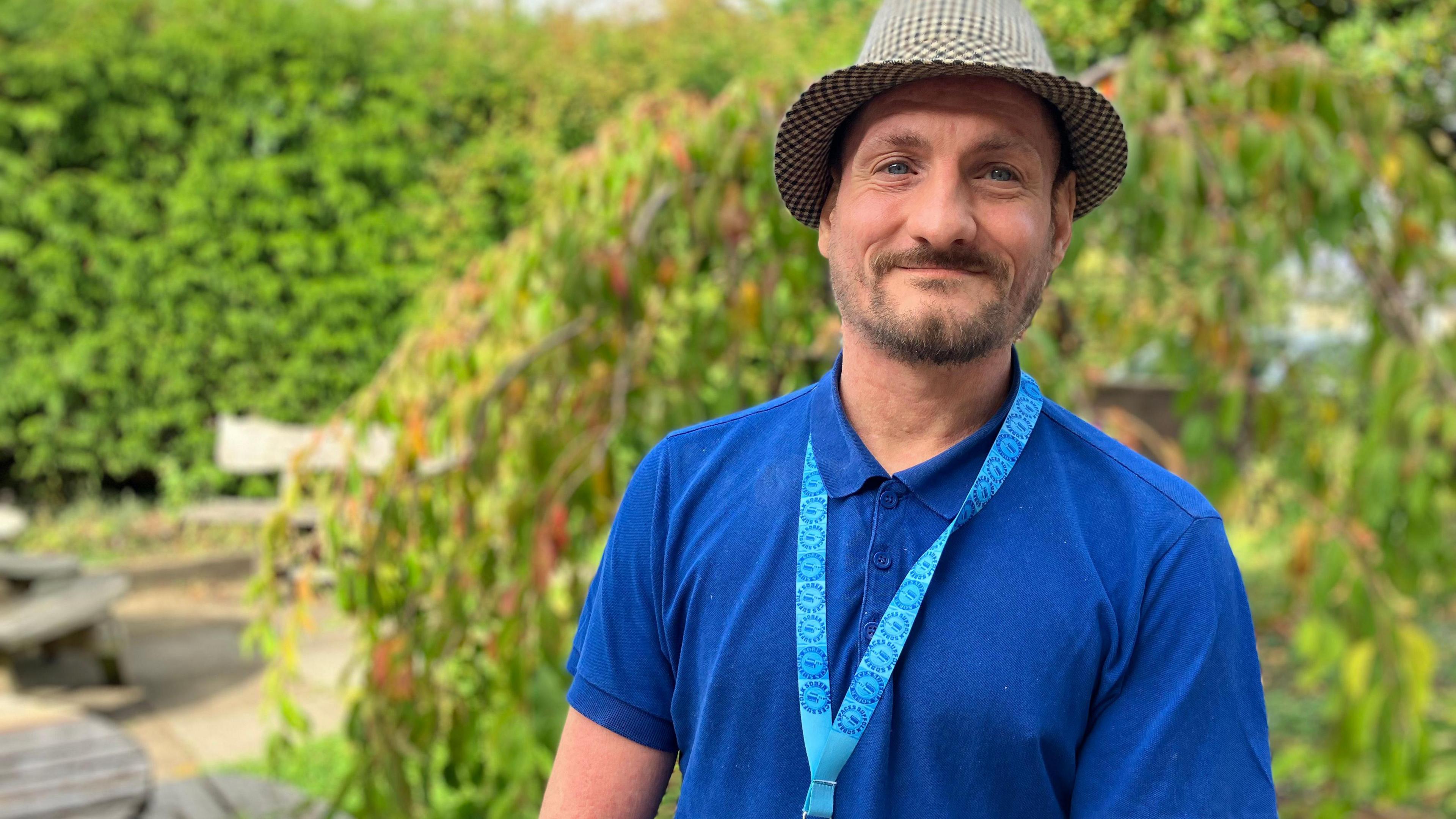 Darryl Cook in a blue top and a hat, with a patterned lanyard round his neck, stands in front of green bushes, smiling at the camera.