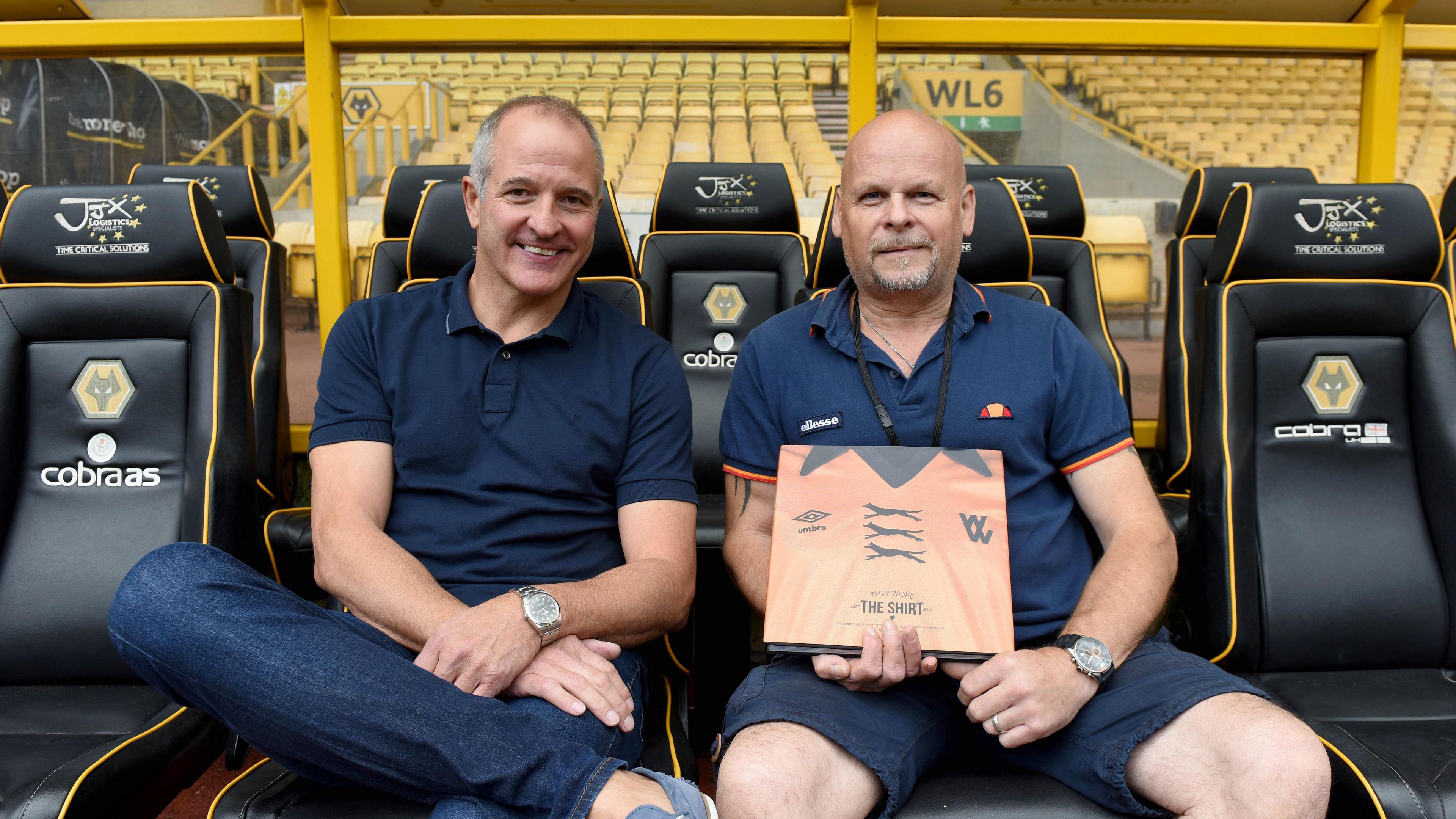 Two men sat in the dugout at a football stadium with a copy of a book about football shirts