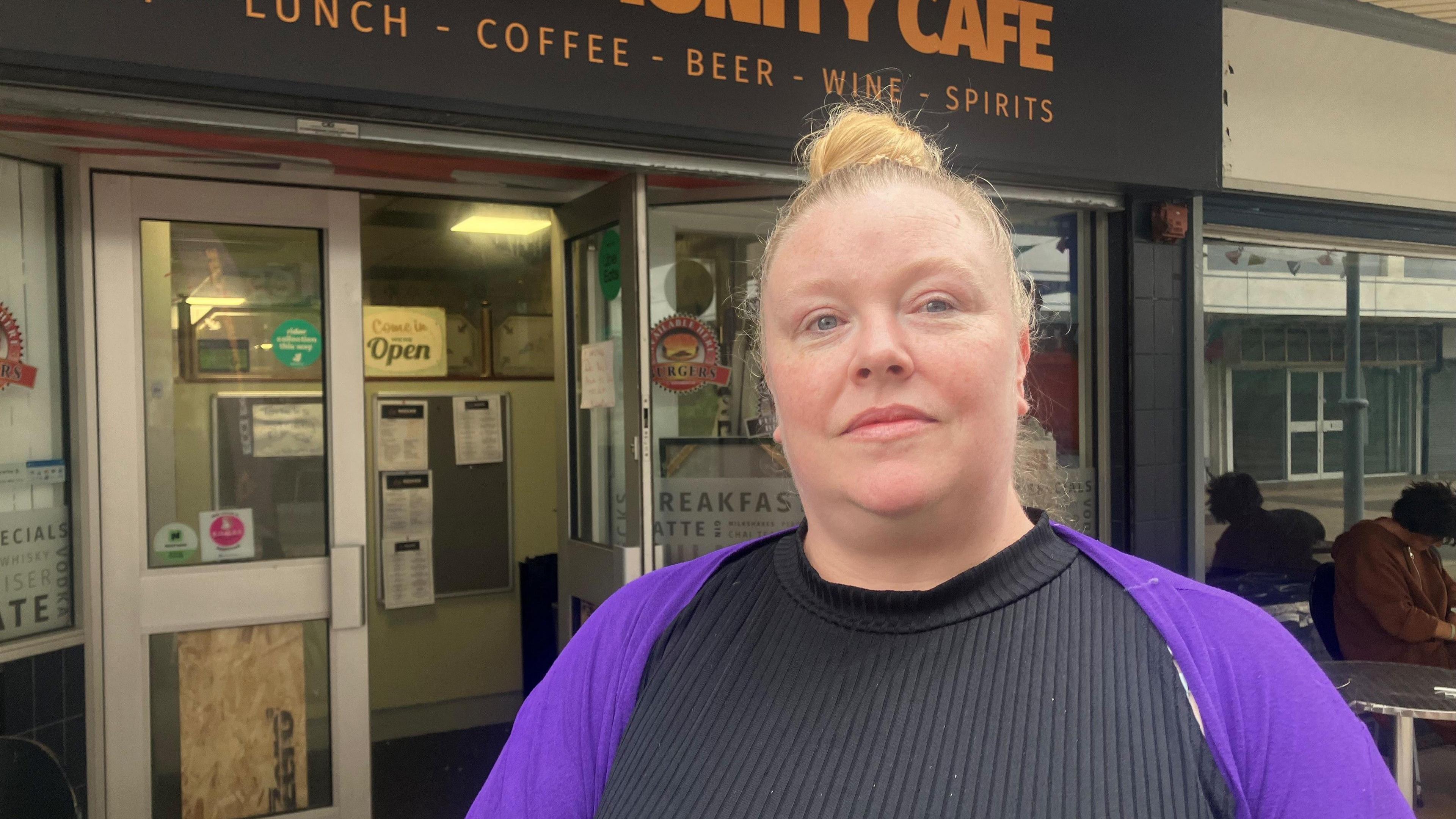 Vicki Warren stands in front of her cafe, one of the remaining open businesses in the shopping centre. She wears a black, ribbed top and a purple cardigan and has blonde hair scraped back. In the background you can see her cafe, with a sign saying it is open. One door is open while the other door has a plywood across the bottom panel of glass.