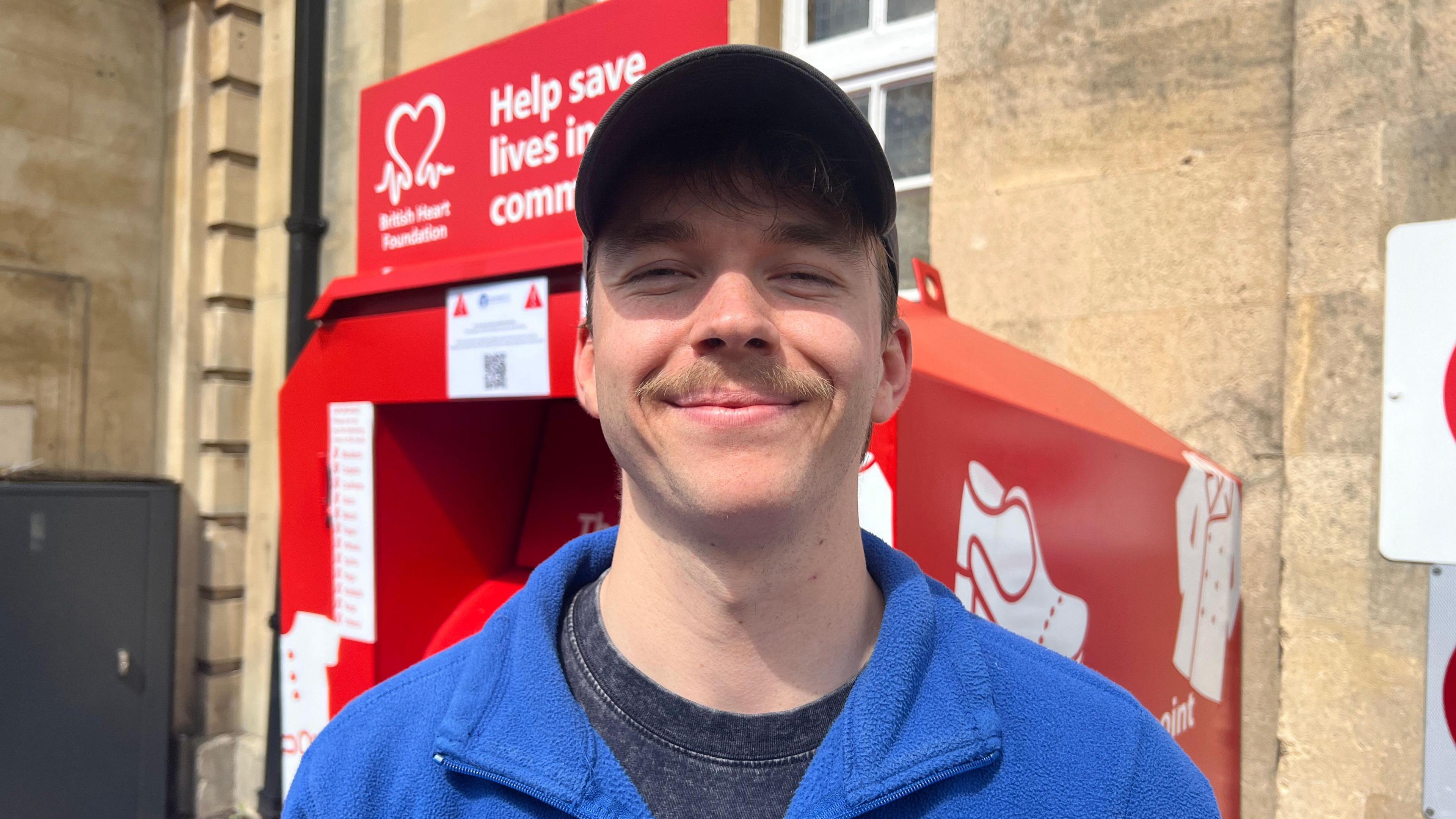 Man in blue zip top wearing a baseball hat, smiling and standing in front of a red British Heart Foundation donation bin