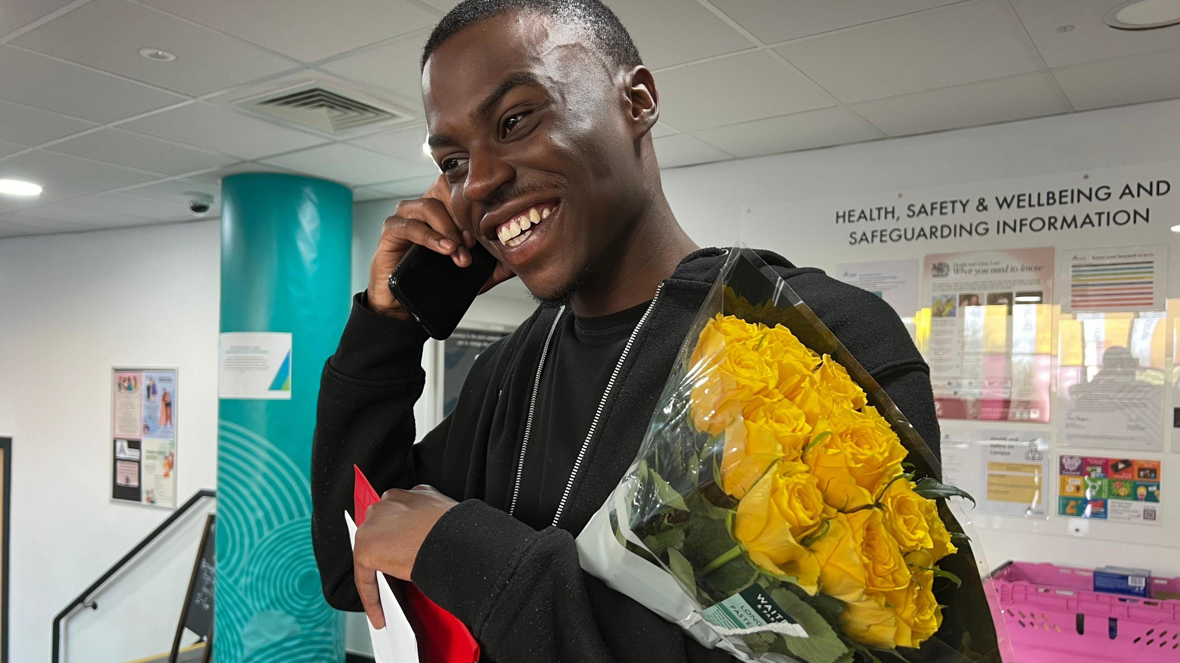 A student is grinning widely, looking away from the camera while he talks on the phone. He wears a long sleeved black hoodie with a silver zip and holds a bunch of yellow roses in the crook of his arm. In the background is a part of the college, there is a blue pillar and a noticeboard behind him.