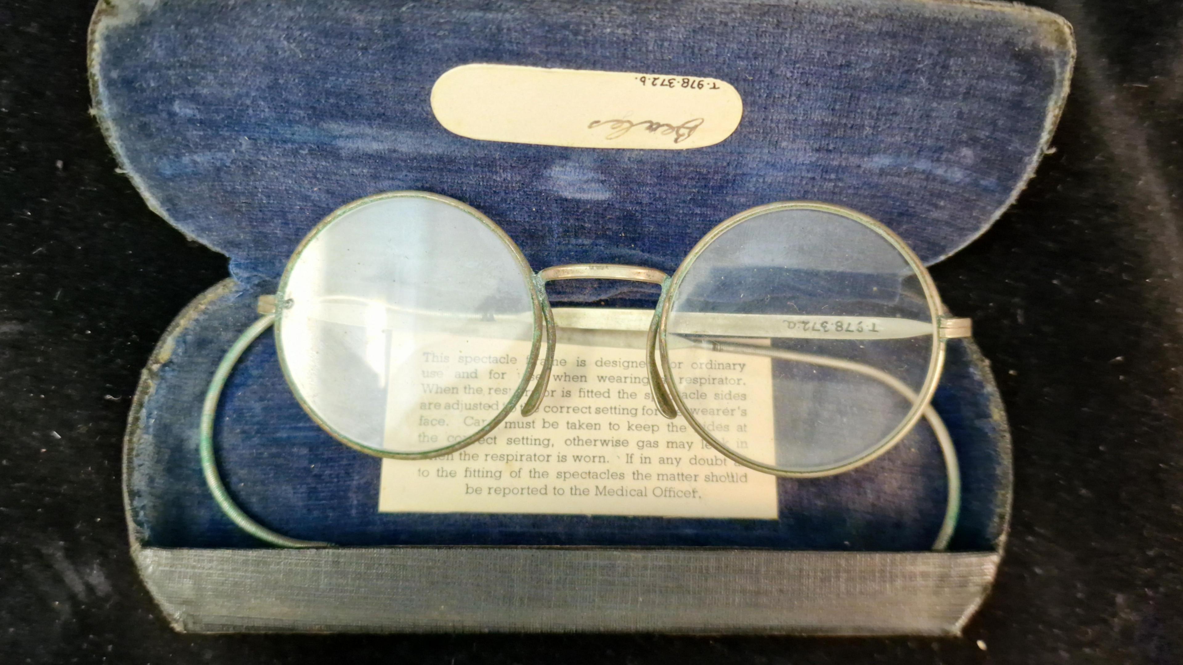 A pair of flat and round rimmed spectacles in a faded blue case with a note underneath saying how they could be worn with a gas mask.