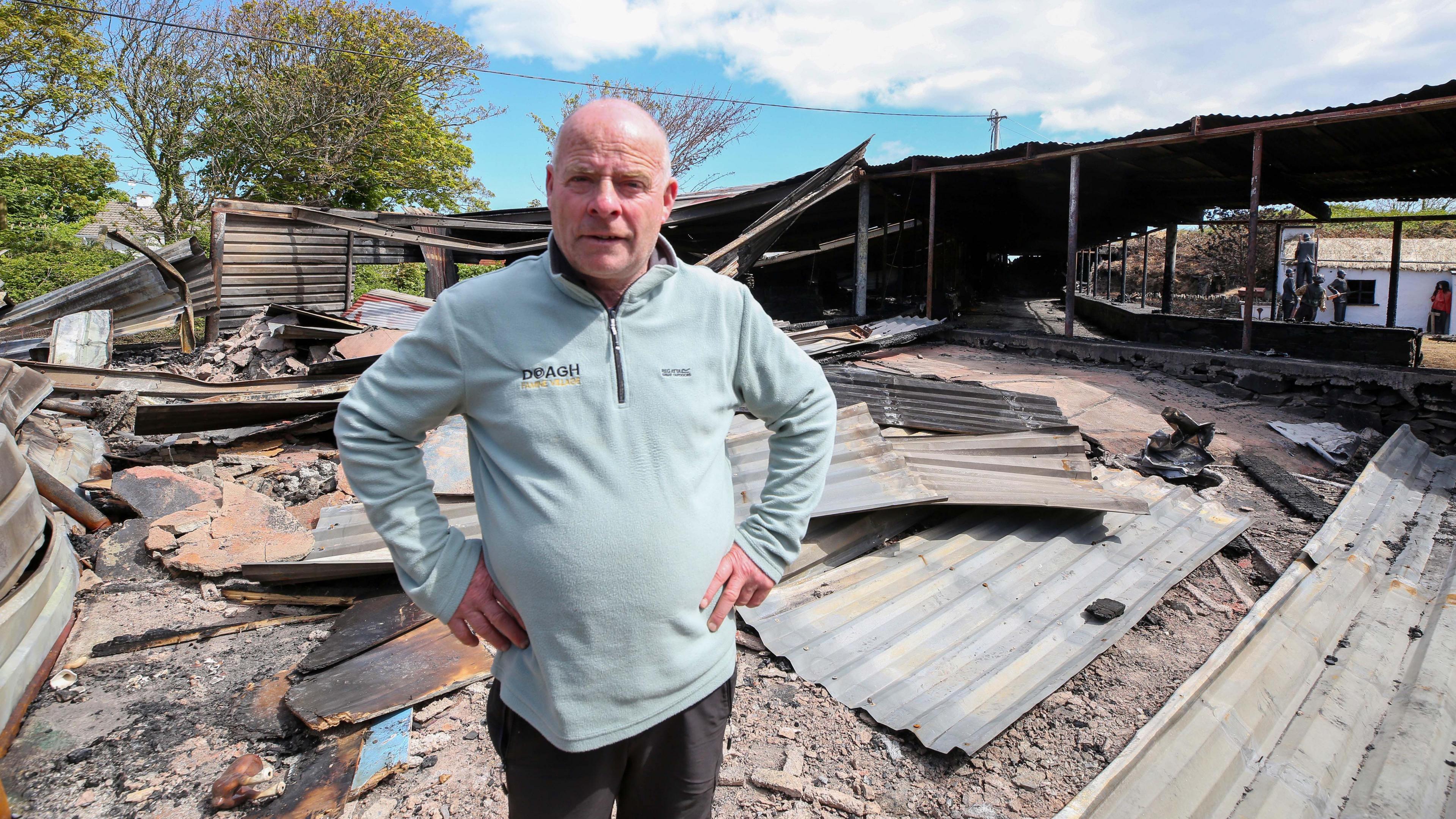 A man wearing a blue jumper stands in the wreckage of a fire. he has his hands on his hips and stares into the camera. There is metal sheeting lying on the ground.