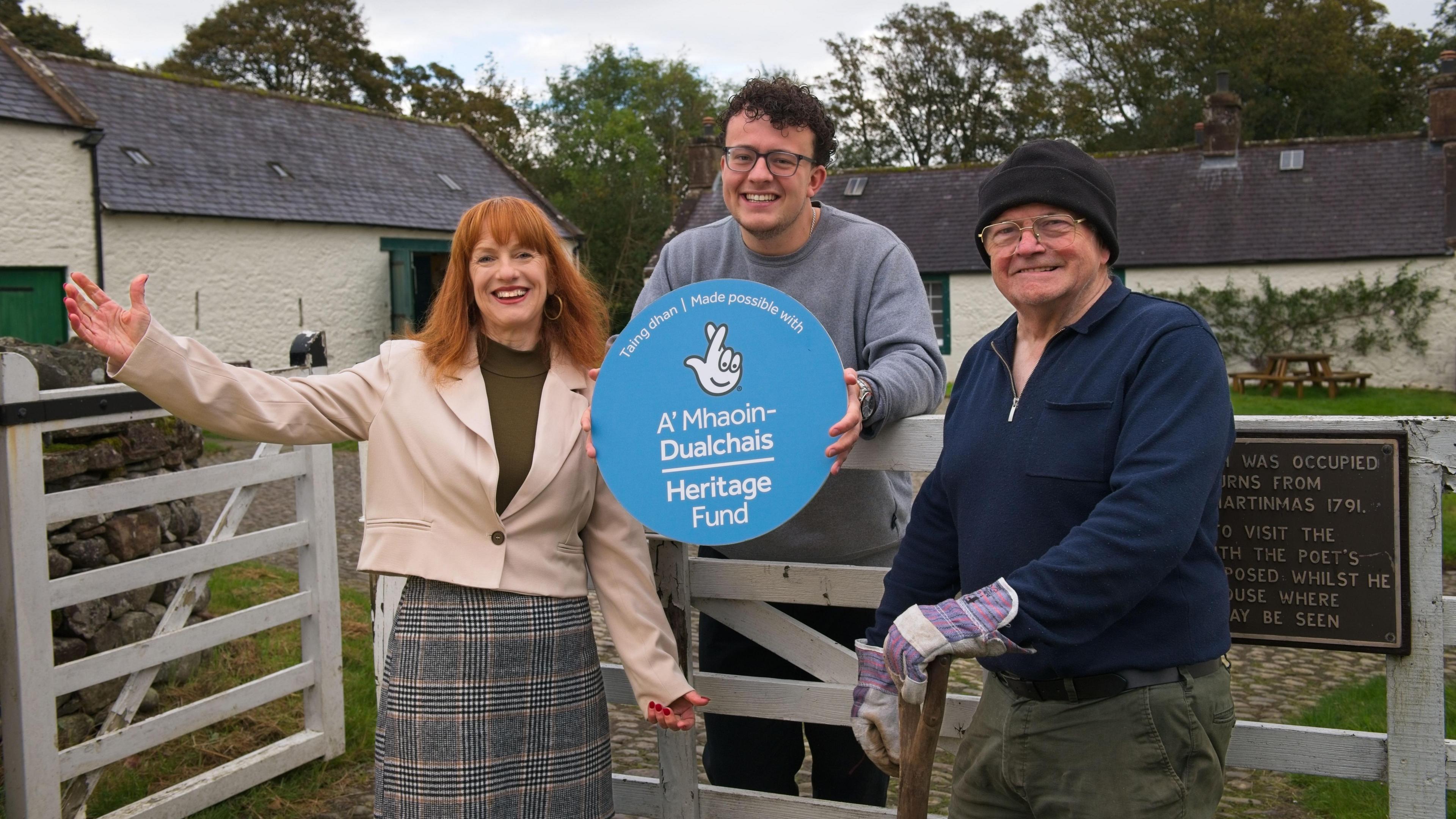 Three people - a red haired woman, a man in glasses and a man in a woolly hat with gardening gloves celebrate money being awarded at Ellisland farm