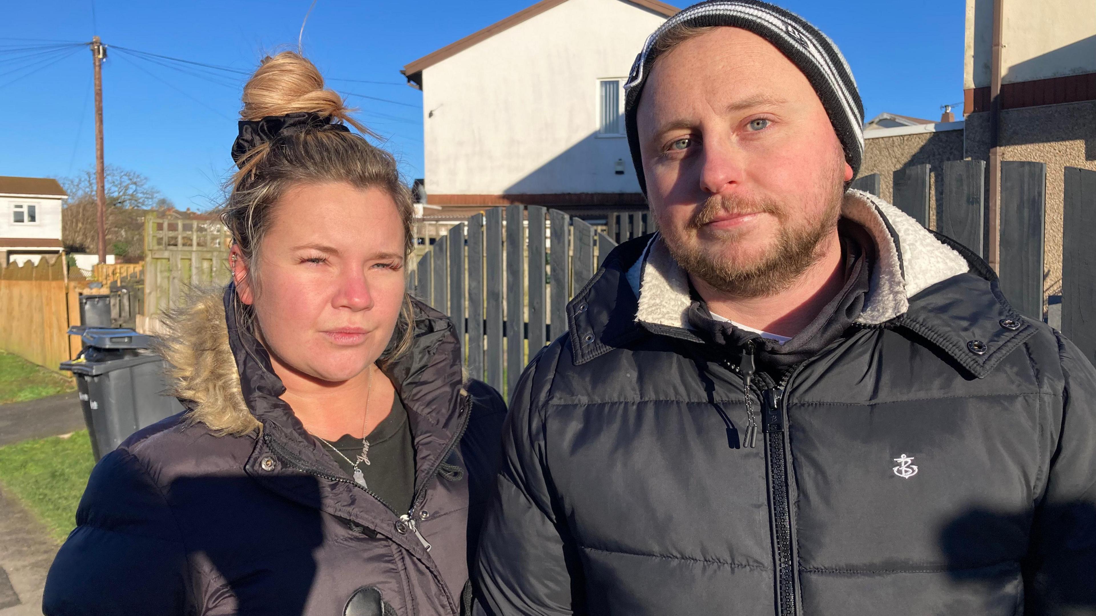 Jenn and Chris Wiley - a couple in their 30s both wearing black coats, stood outside a house with a fence and wheelie bin in the background