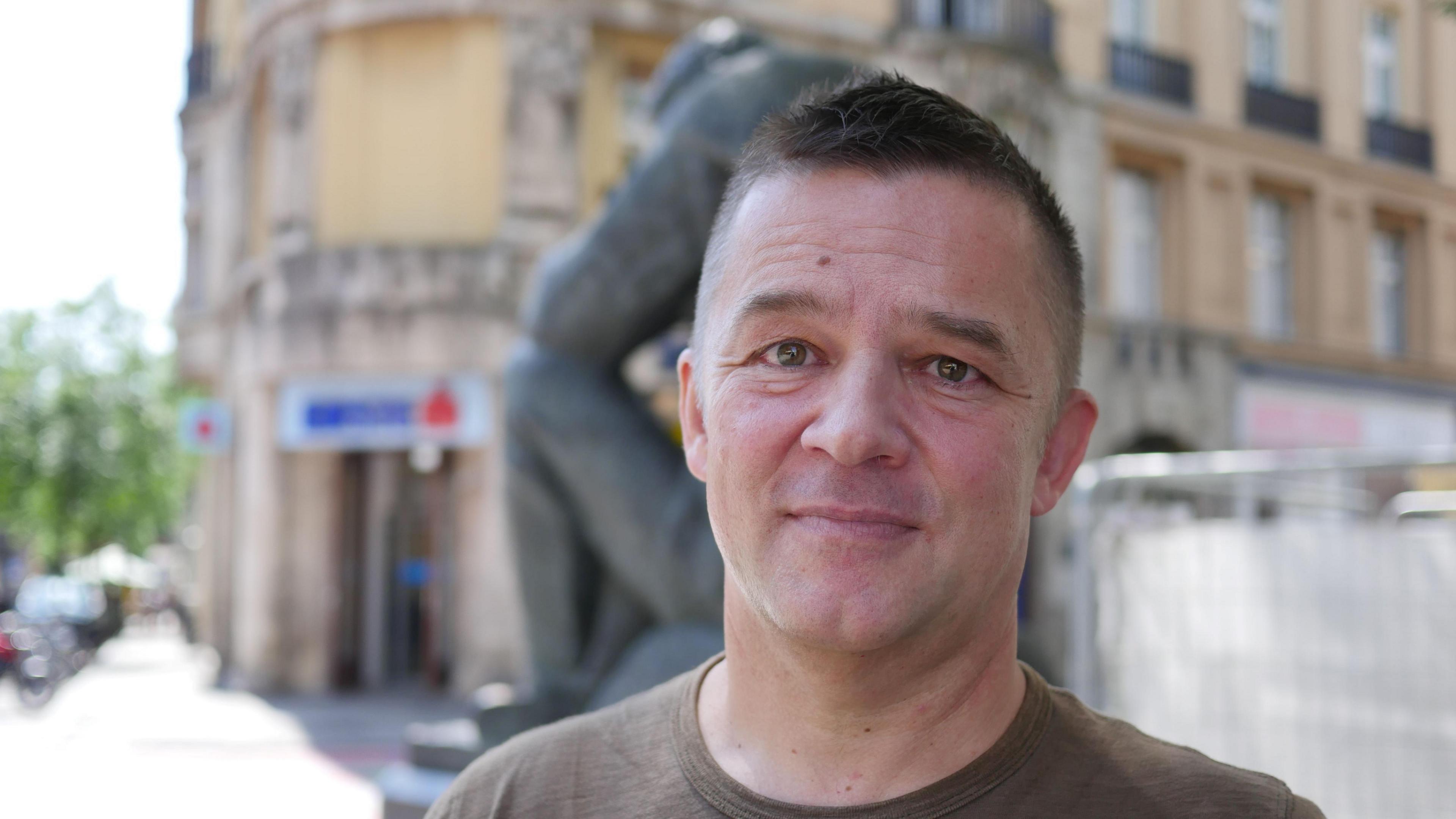 Tvrtko Jakovina looks into the camera smiling faintly. He has short dark hair and is wearing a khaki t-shit. He is standing on the pavement of a quiet street.