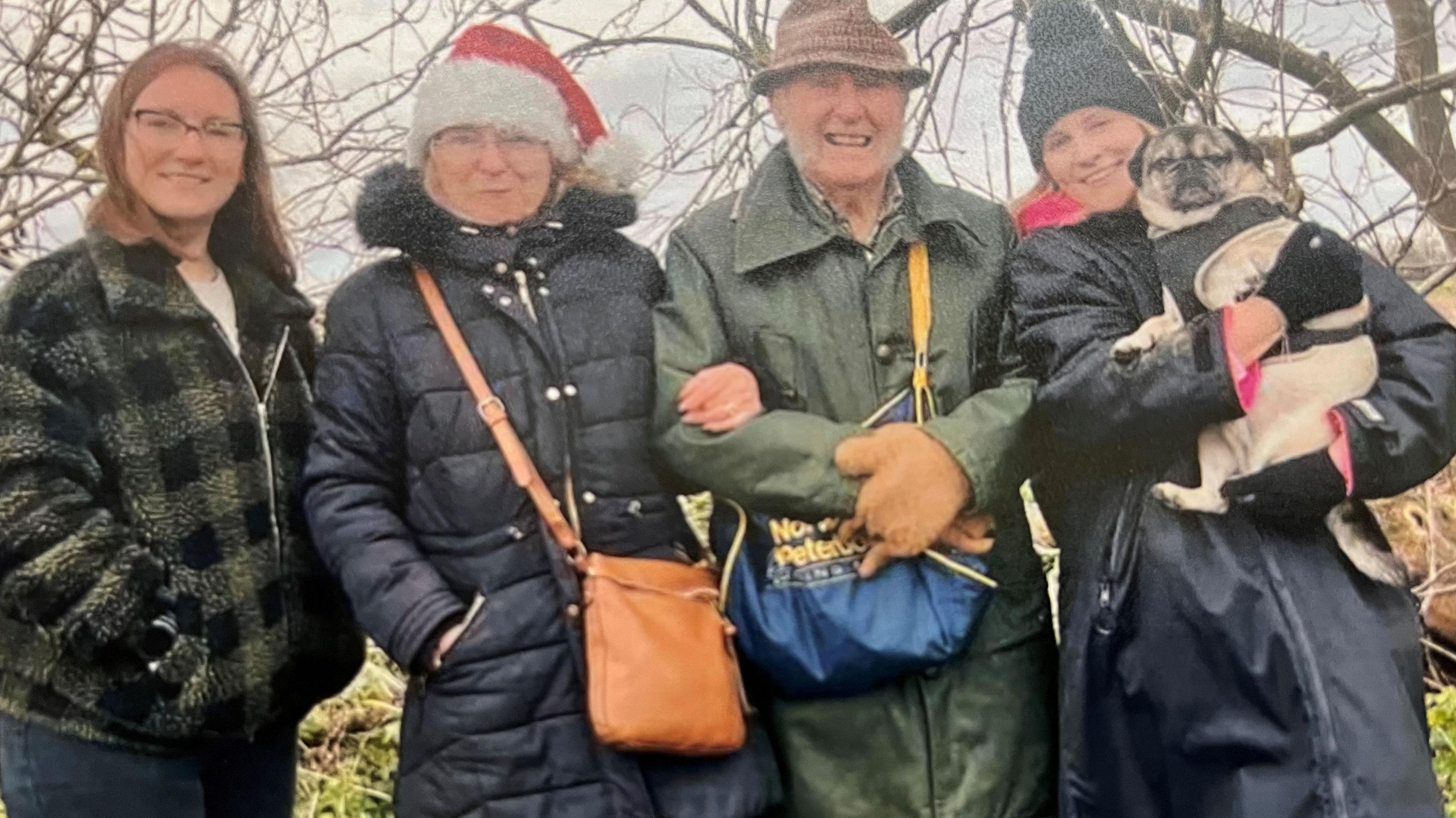 Colin Bedford, 88, wearing a green wax jacket overcoat, he has a tartan fedora hat and long grey whiskery sideburns, and has light coloured gloves,
he is flanked by his grand-daughters and an older lady who has a red Santa hat, that is his daughter, who went on the very first walk, aged one. In the background are tree branches and farmland.