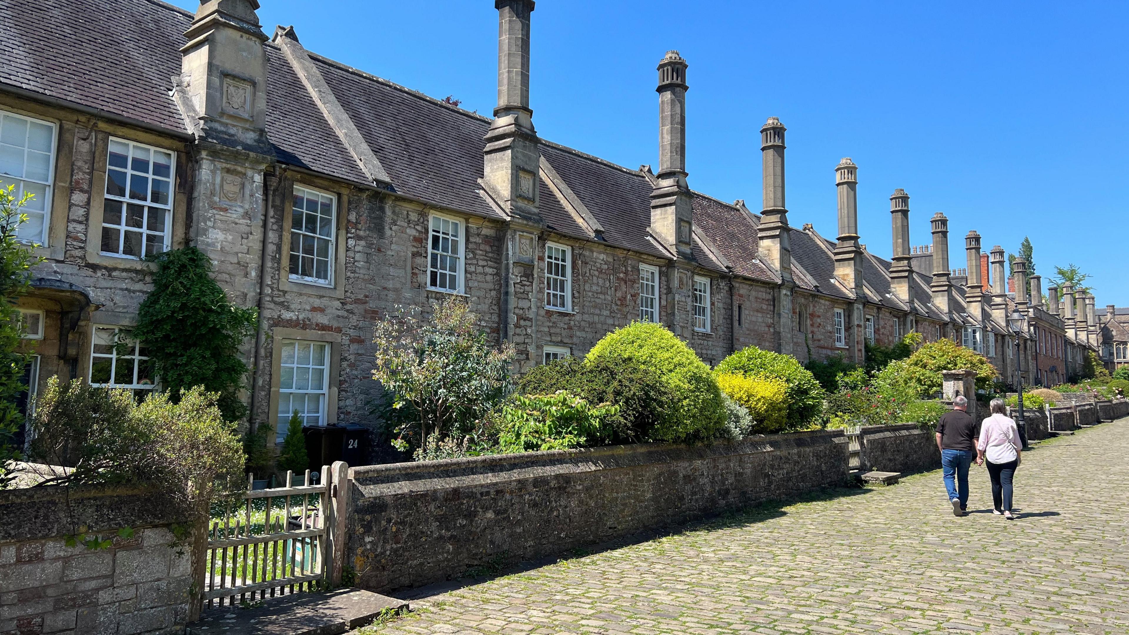 A row of old stone terraced houses with tall chimneys and a cobbled street outside. The houses have walled front gardens, which have green bushes and trees inside. It is a sunny day and there is a man and woman walking up the cobbled street holding hands. 