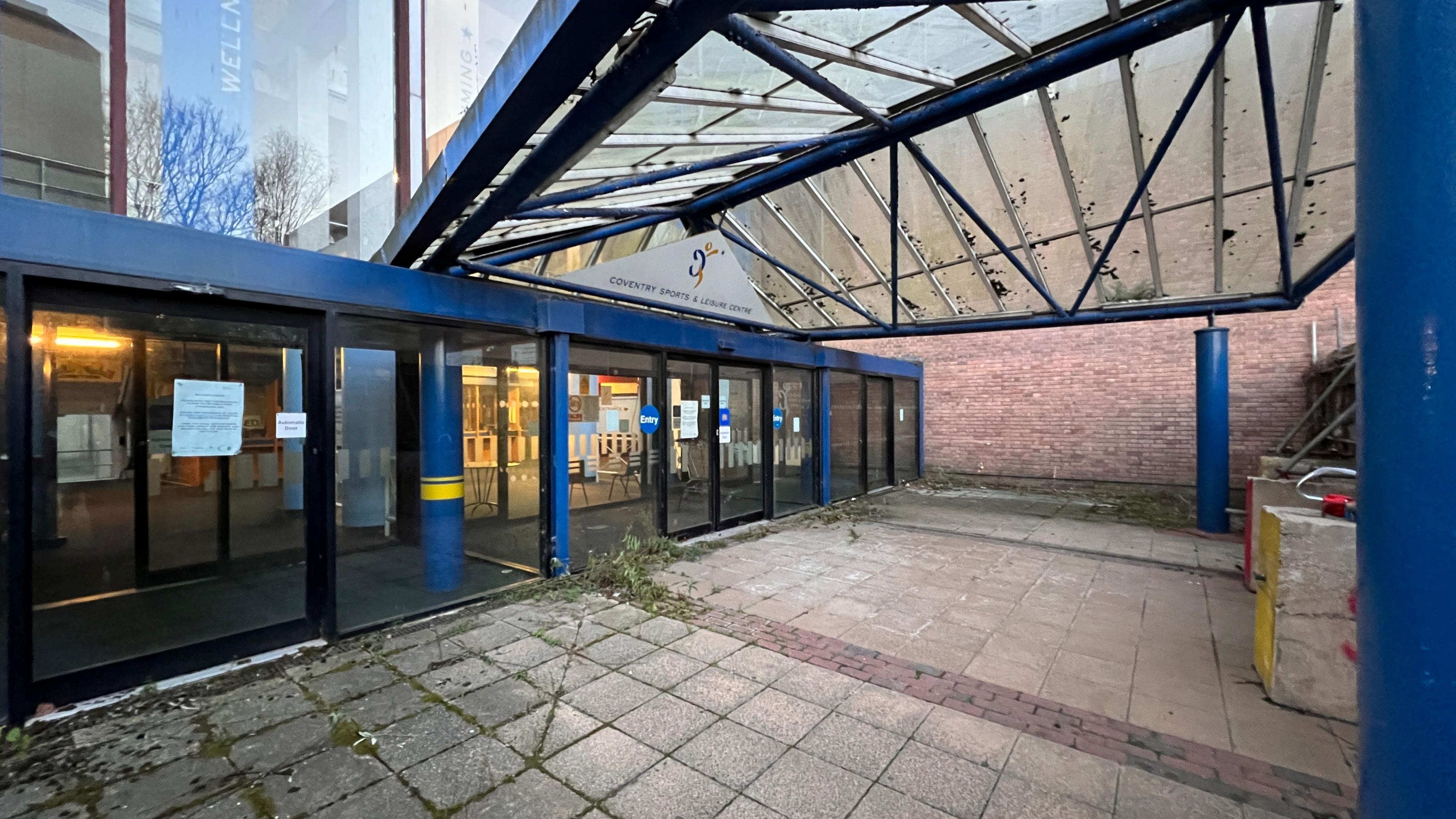 Image shows the entrance to Coventry Sports and Leisure Centre, in Fairfax Street. The door surrounds and pillars are pained blue, a transparent canopy is dirty and covered in moss. Weeds protrude from the paving slabs leading to the sliding doors.