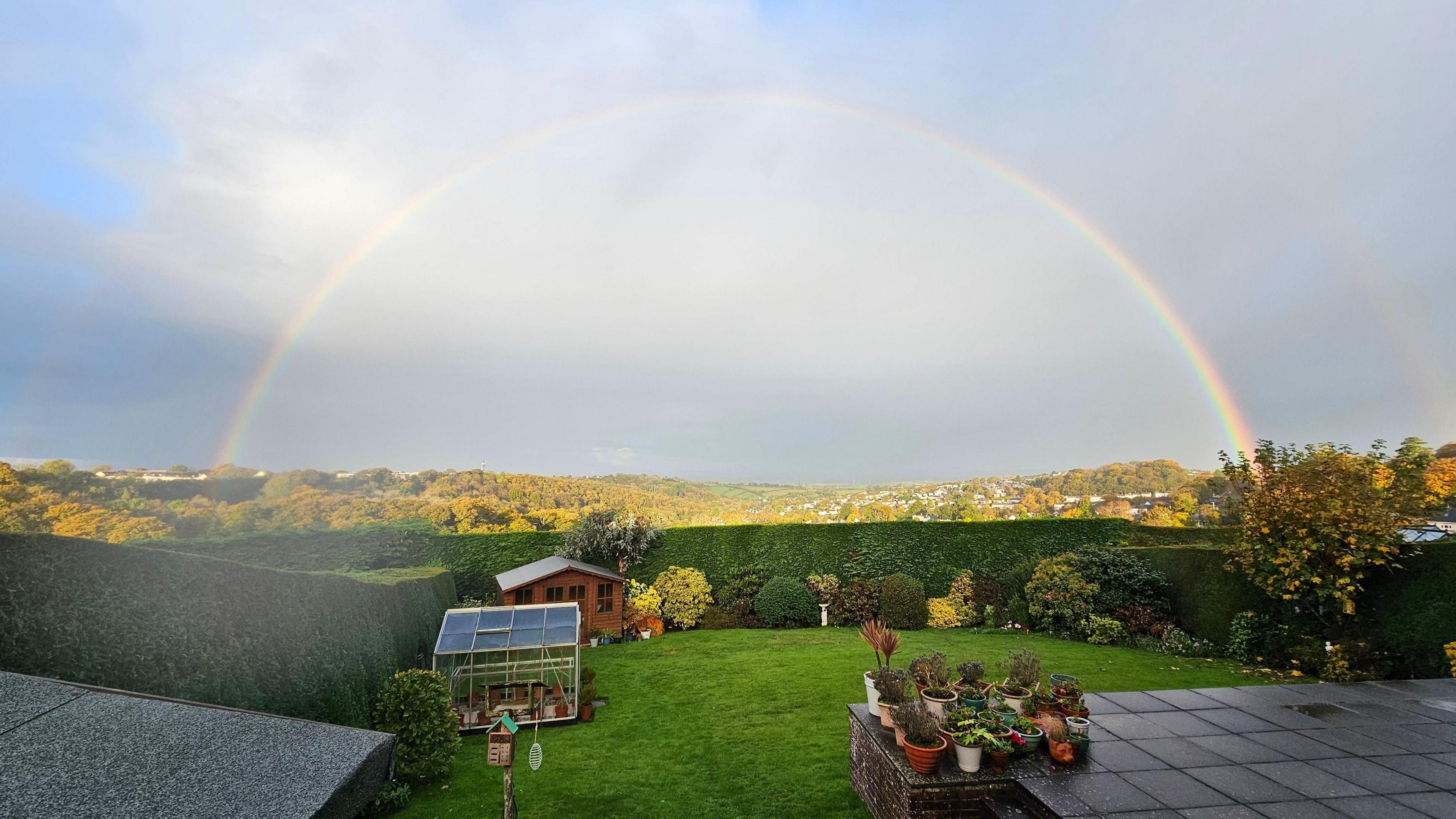 A rainbow semi-circle. The sky is filled with grey cloud and some of the sky is light blue. The picture is taken from the back garden of a house. A green lawn can be seen with shrubbery and there is a garden shed and a greenhouse to the left of the garden with a patio to the right.